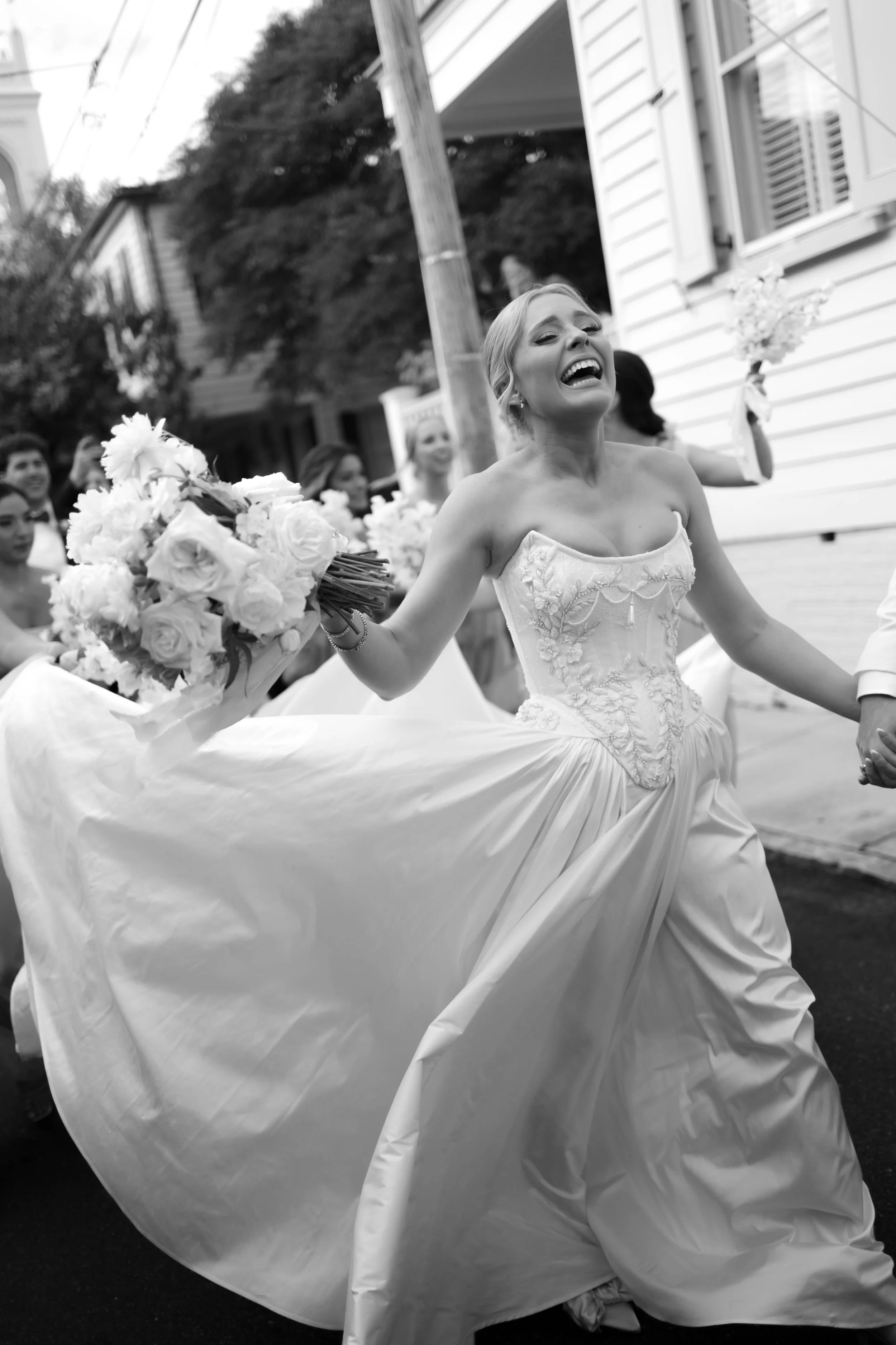 A joyful bride in a strapless wedding gown holding a bouquet of flowers, walking in a parade or celebration with friends and family in a neighborhood street.