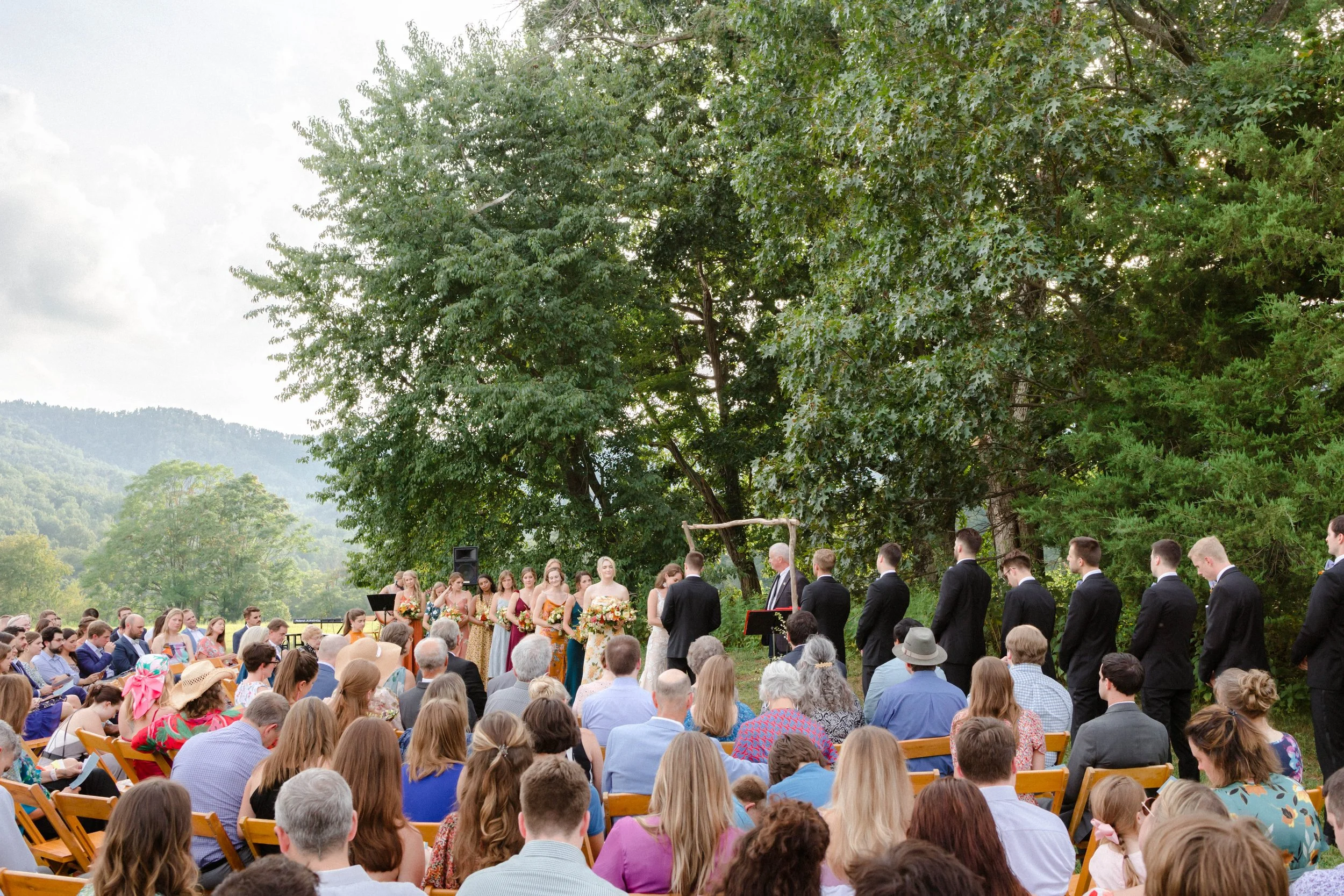 Outdoor wedding ceremony with guests seated on wooden chairs, a bride and groom at an arch, surrounded by bridesmaids and groomsmen, under large trees and scenic mountains.