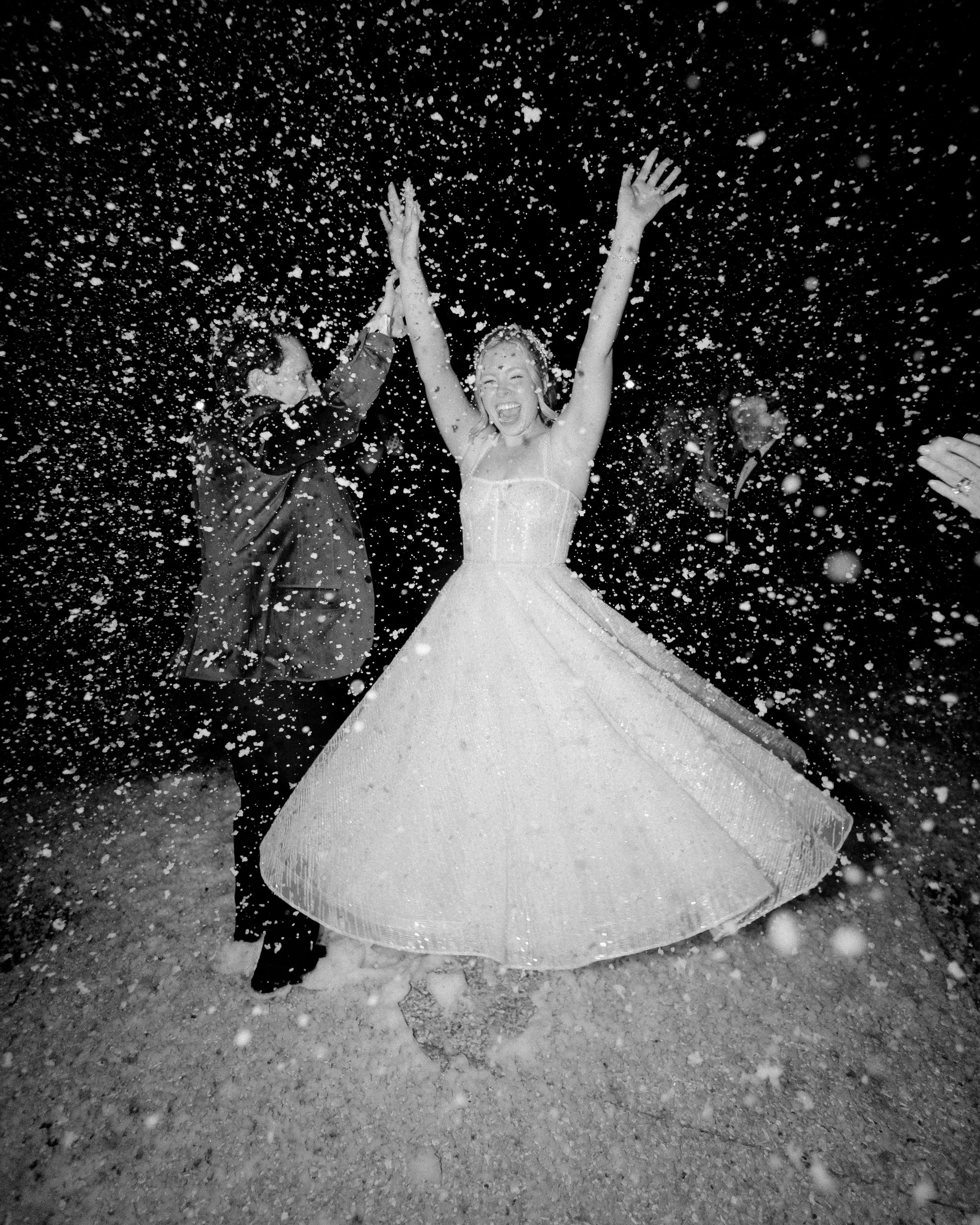 Black and white photo of a woman in a wedding dress celebrating with her arms raised while a man claps next to her, surrounded by falling snow or confetti.