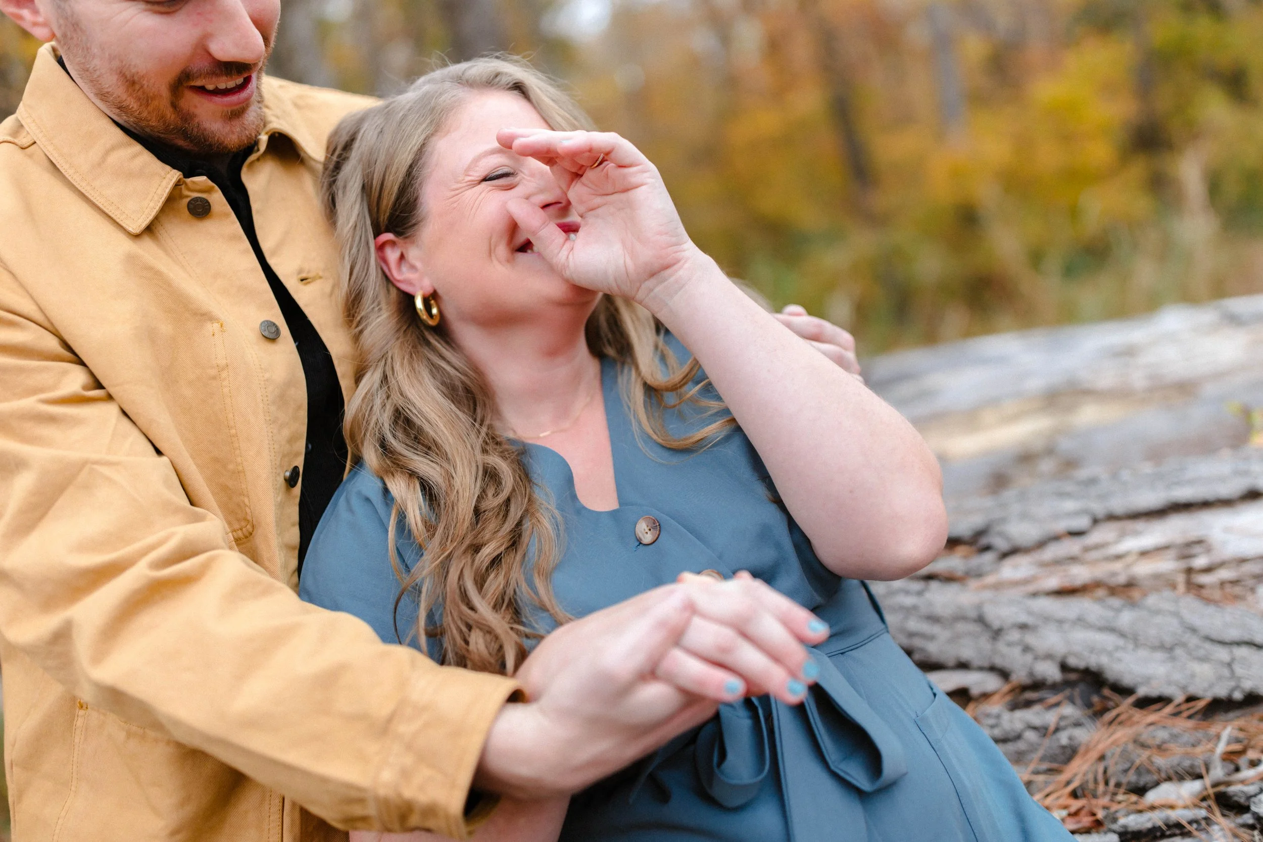 A man and woman outdoors smiling and laughing, with autumn trees and rocks in the background. The woman is covering her eyes with her hand and smiling, while the man holds her by the waist.