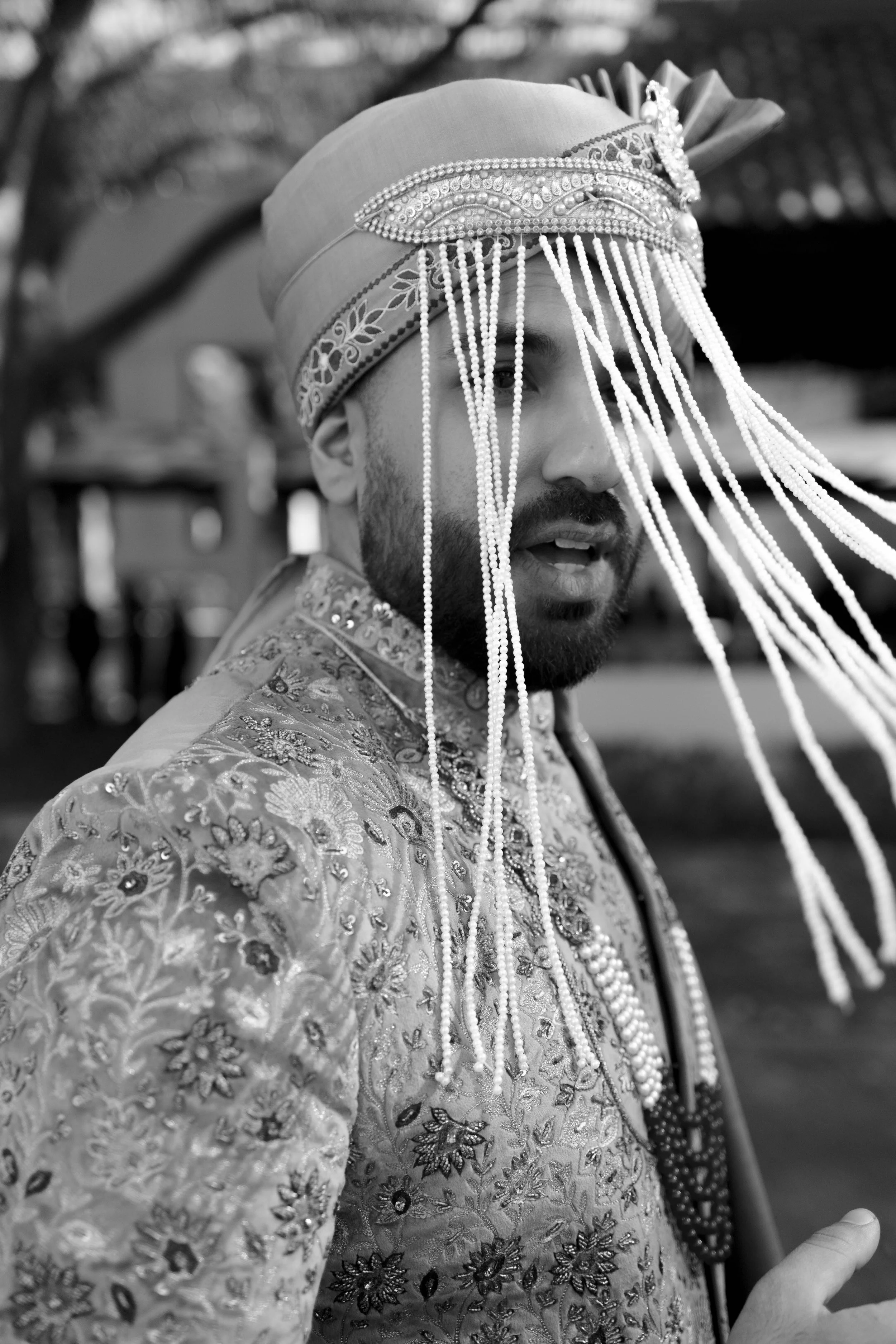 A man dressed in traditional Indian wedding attire, wearing ornate embroidery and jewelry, with a headpiece covering part of his face.