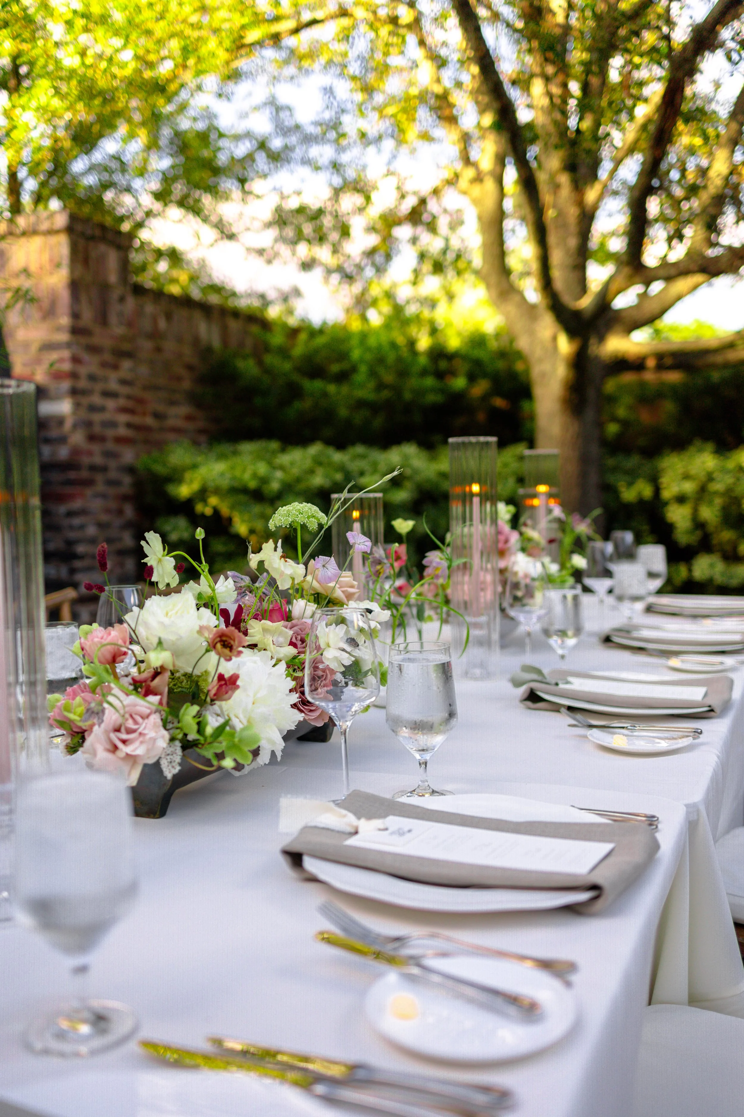 A beautifully set outdoor dining table decorated with flowers, glasses, and candles under a large tree.