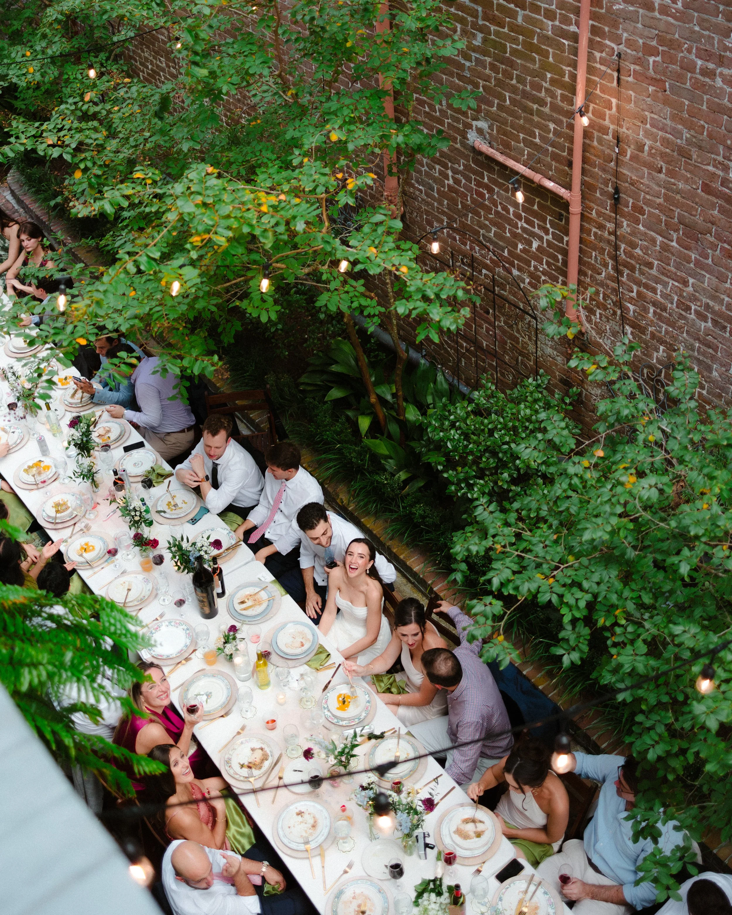 People sitting at long dining table outdoors during a celebration or dinner event, with greenery and string lights overhead.