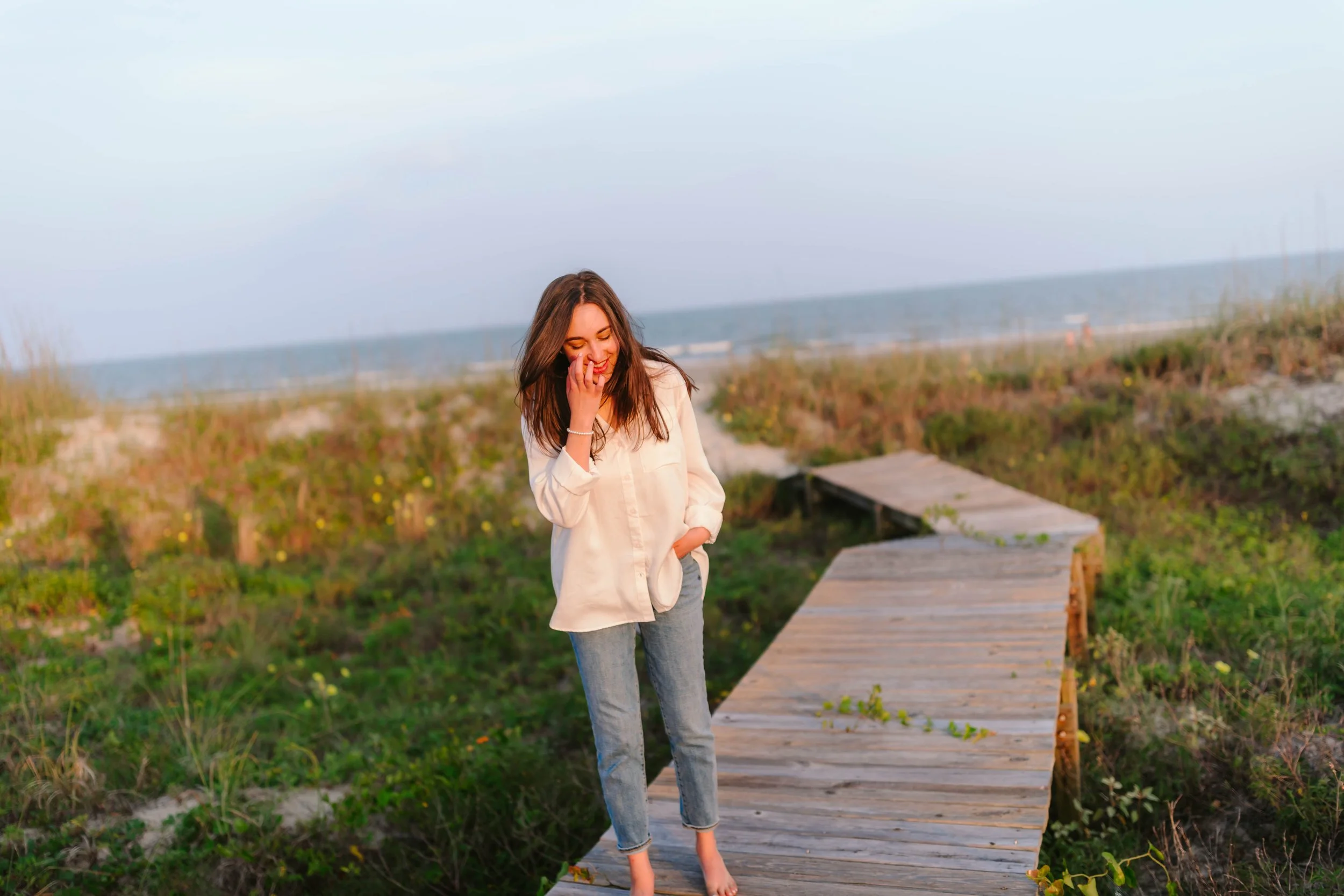 A woman walking barefoot on a wooden boardwalk at the beach, smiling and covering her mouth with her hand, with ocean and cloudy sky in the background.