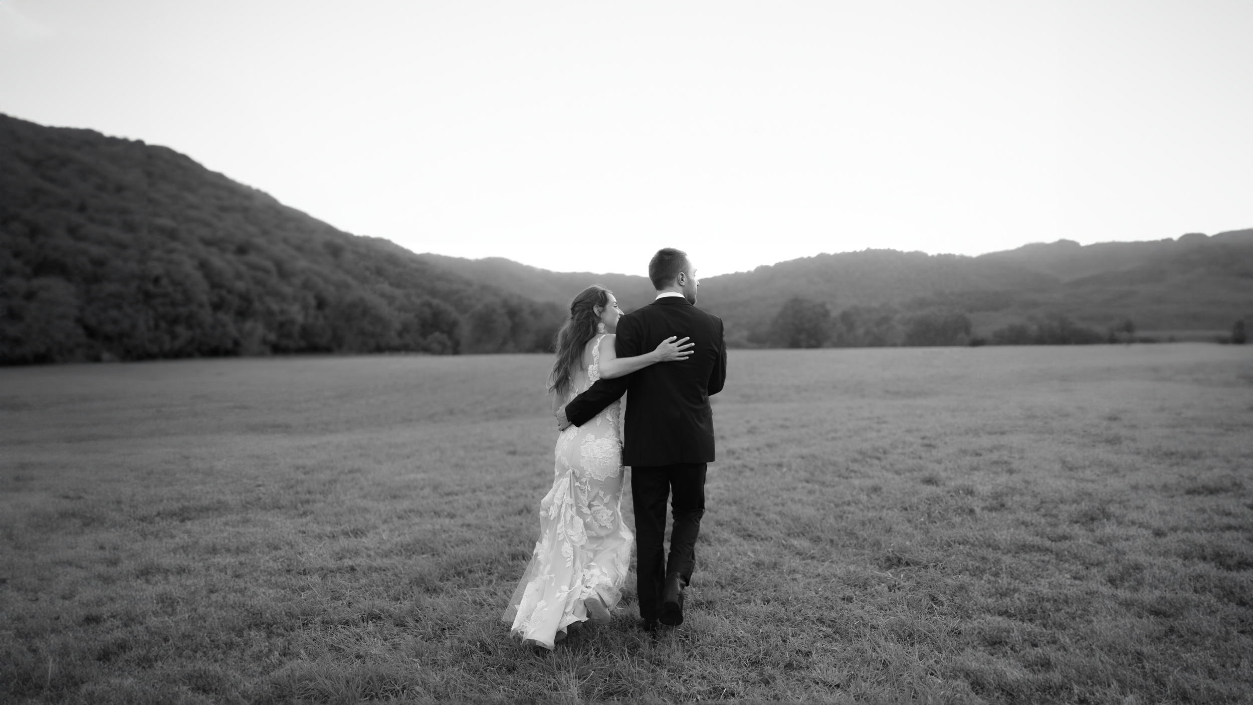A bride and groom walking hand in hand across an open field with hills in the background, captured in black and white.