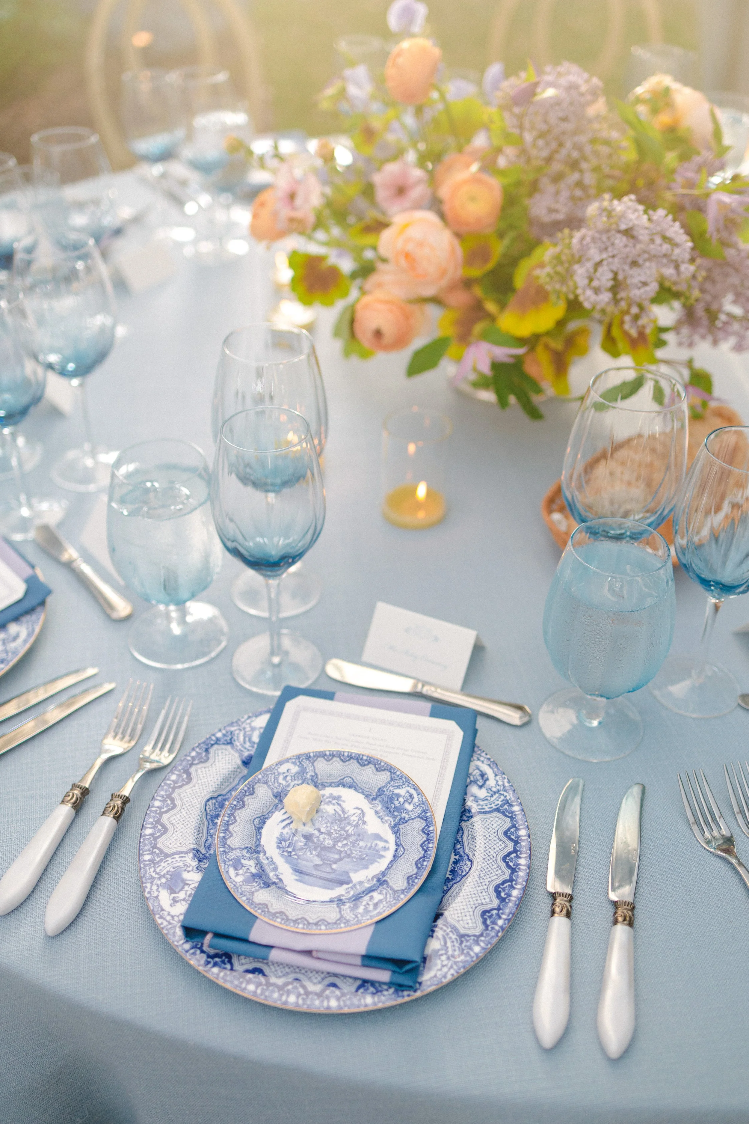 Elegant table setting with blue and white china, silverware, and water glasses, decorated with a floral centerpiece of pastel-colored flowers and a lit candle.