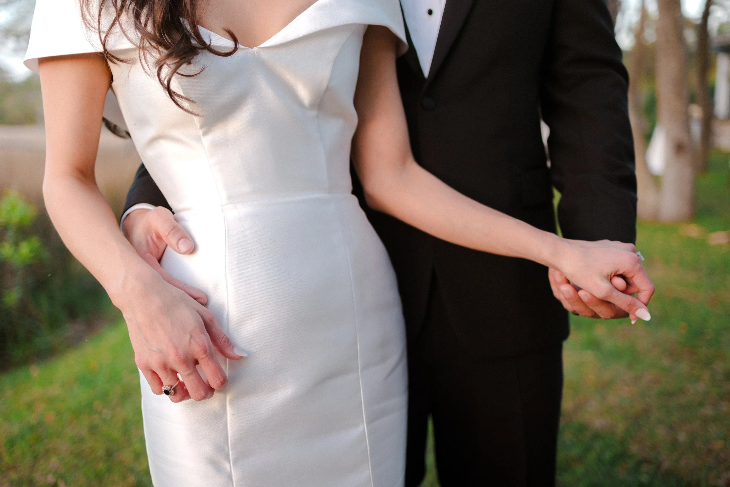 A close-up of a couple holding hands, with the woman wearing a white satin dress and a wedding ring, and the man in a black suit, outdoors with trees and greenery in the background.