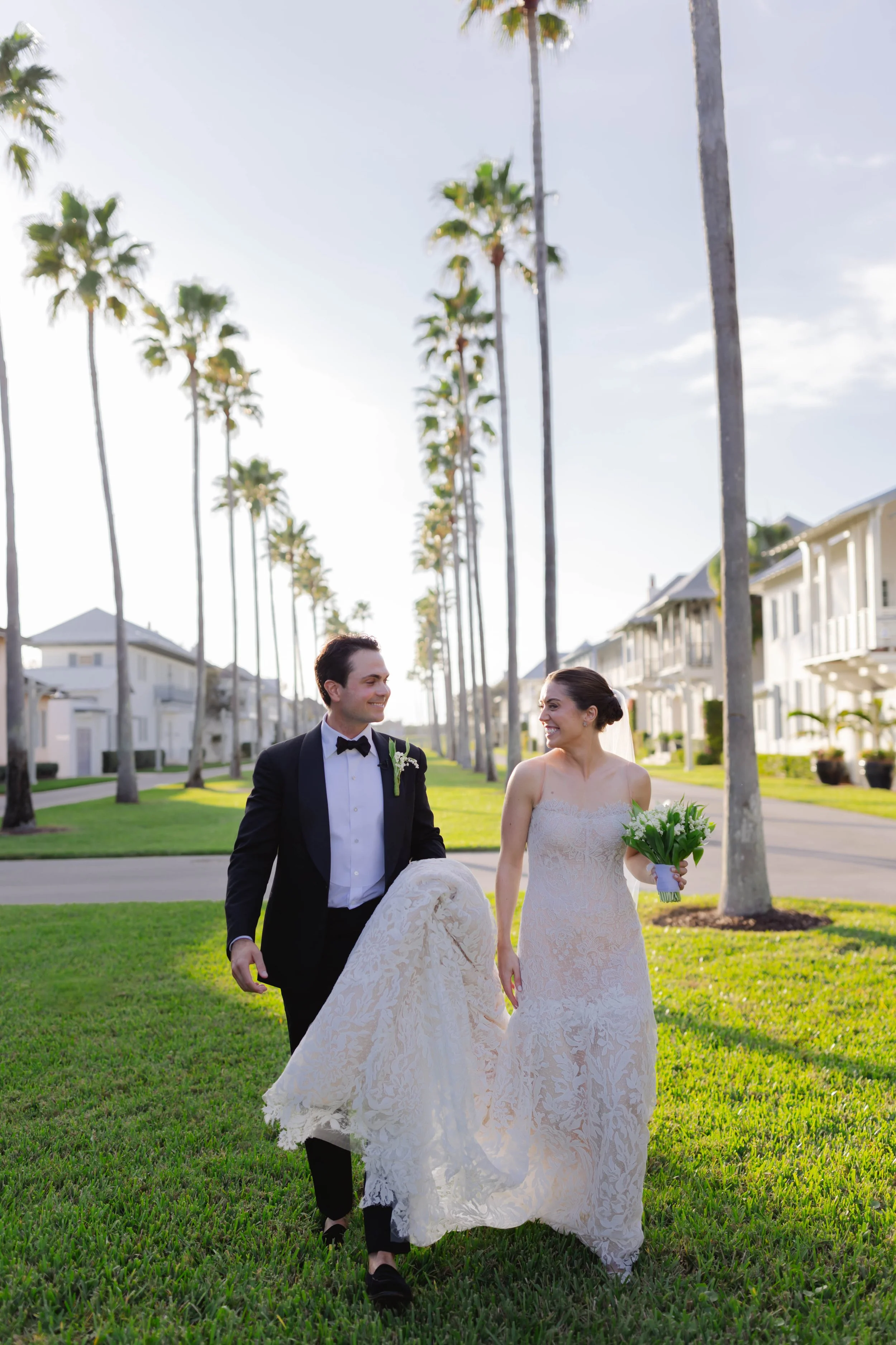 A newlywed couple walking on a grassy lawn, the groom in a tuxedo holding the bride’s train, the bride in a lace wedding dress holding a bouquet, with palm trees and white houses in the background on a sunny day.