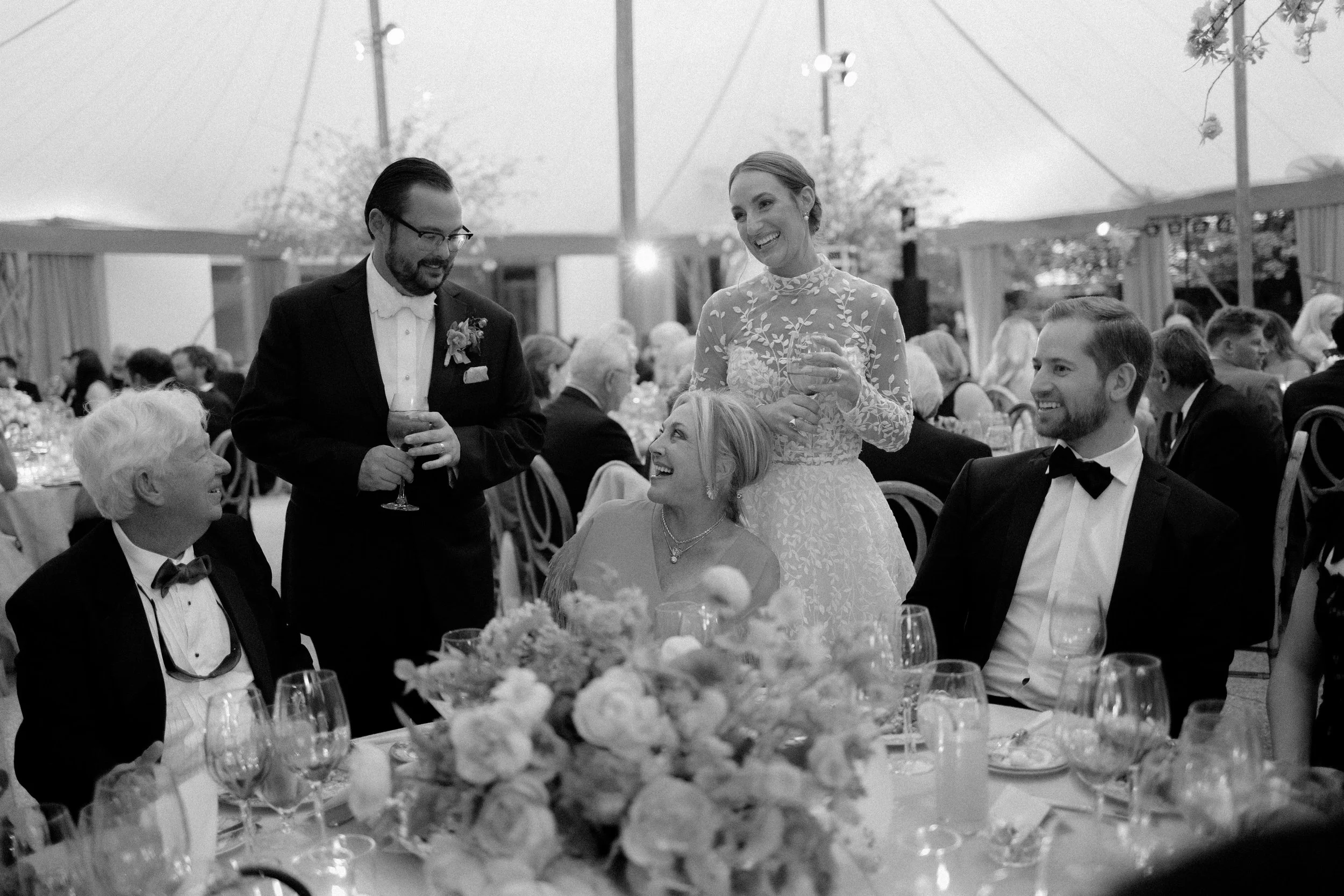 A group of people at a wedding reception, including a woman in a lace dress standing and talking with a glass of wine, a man in a tuxedo holding a drink, and three seated guests engaged in conversation, with a floral centerpiece on the table.
