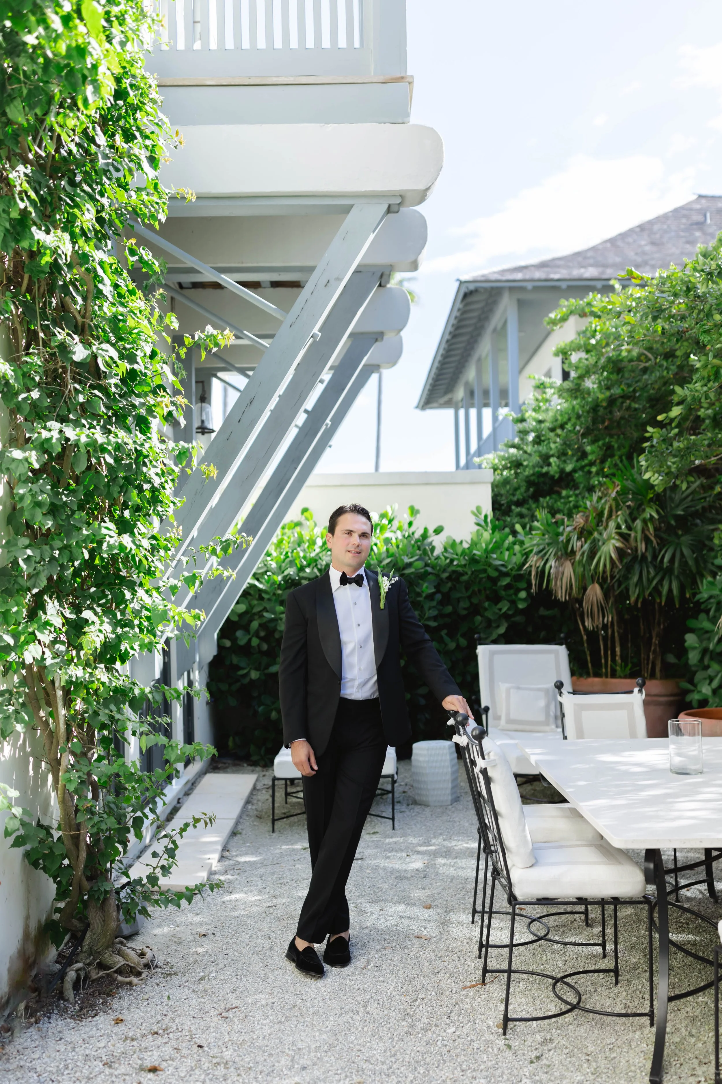 A man in a black tuxedo with a bow tie and boutonniere standing on a patio, holding a chair, with outdoor furniture and lush greenery in the background.