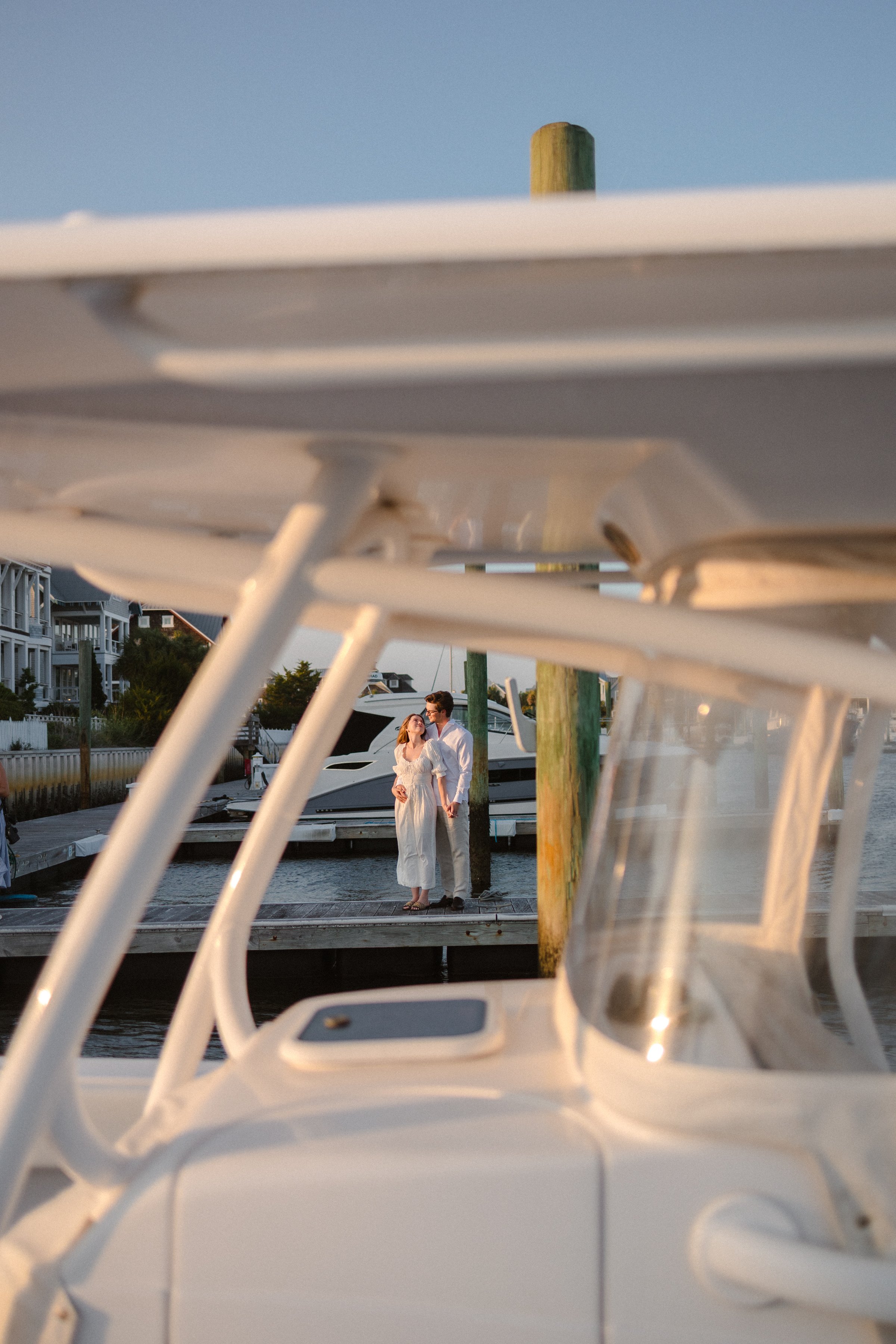 A couple standing on a dock by boats, viewed through a boat's framing, during sunset.