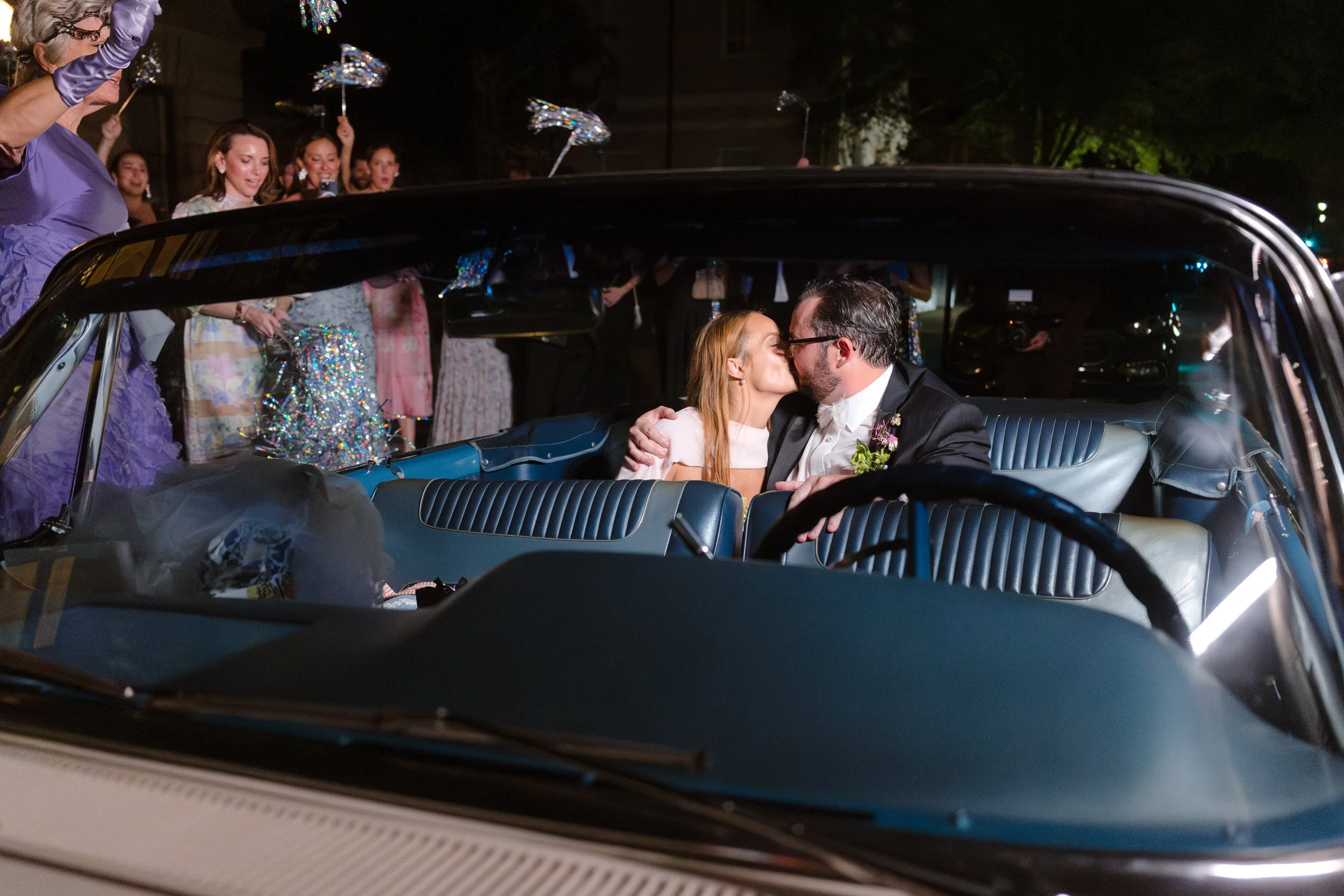 A bride and groom share a kiss inside a vintage car, surrounded by friends and family during the evening wedding celebration.