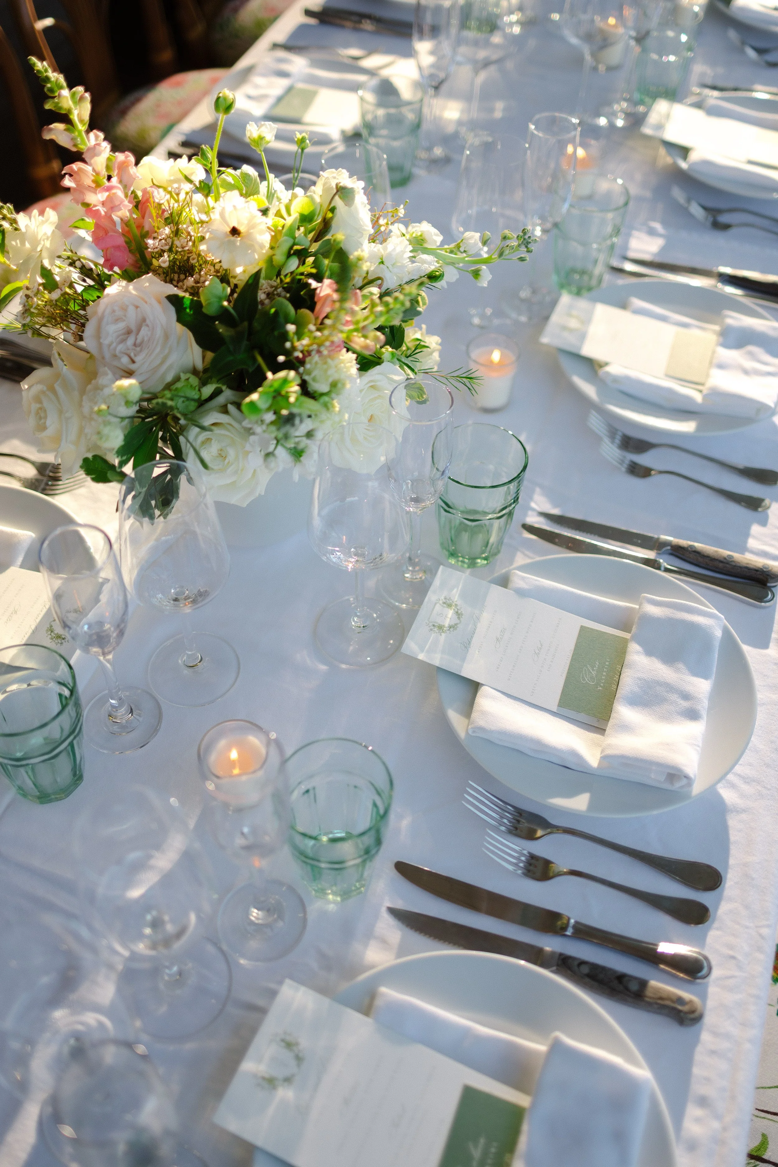 A beautifully set dining table with a white tablecloth, a floral centerpiece with white and pink flowers, multiple glasses, white plates with napkins, and silverware.