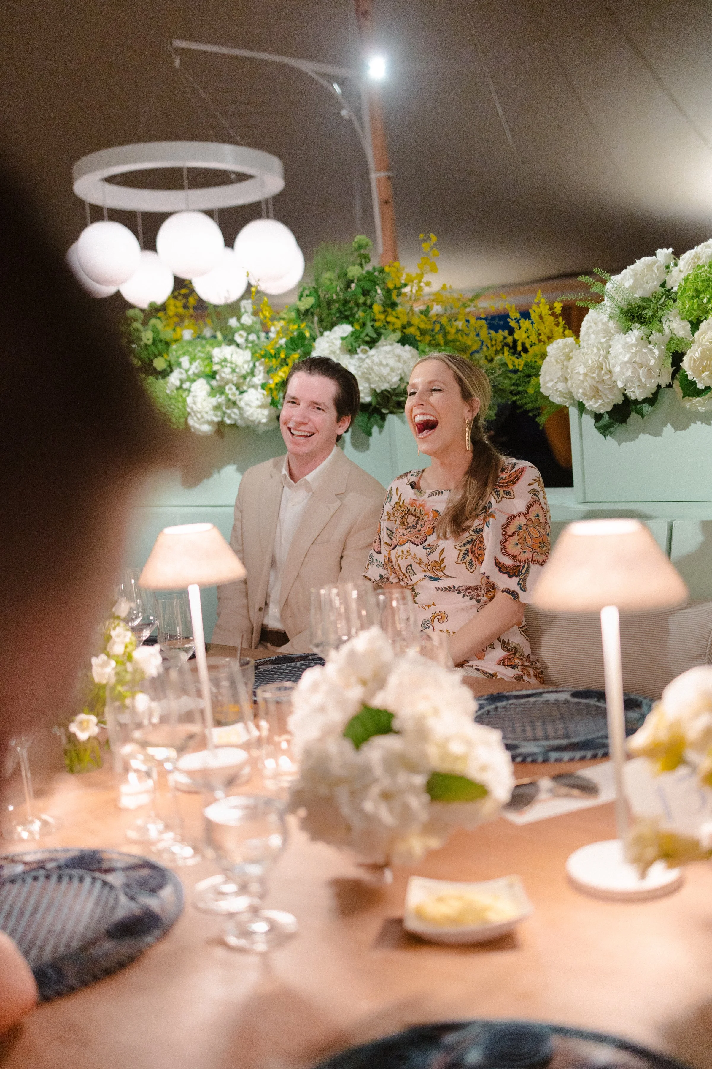 People laughing and enjoying a celebration at a decorated dinner table with white flowers and soft lighting.