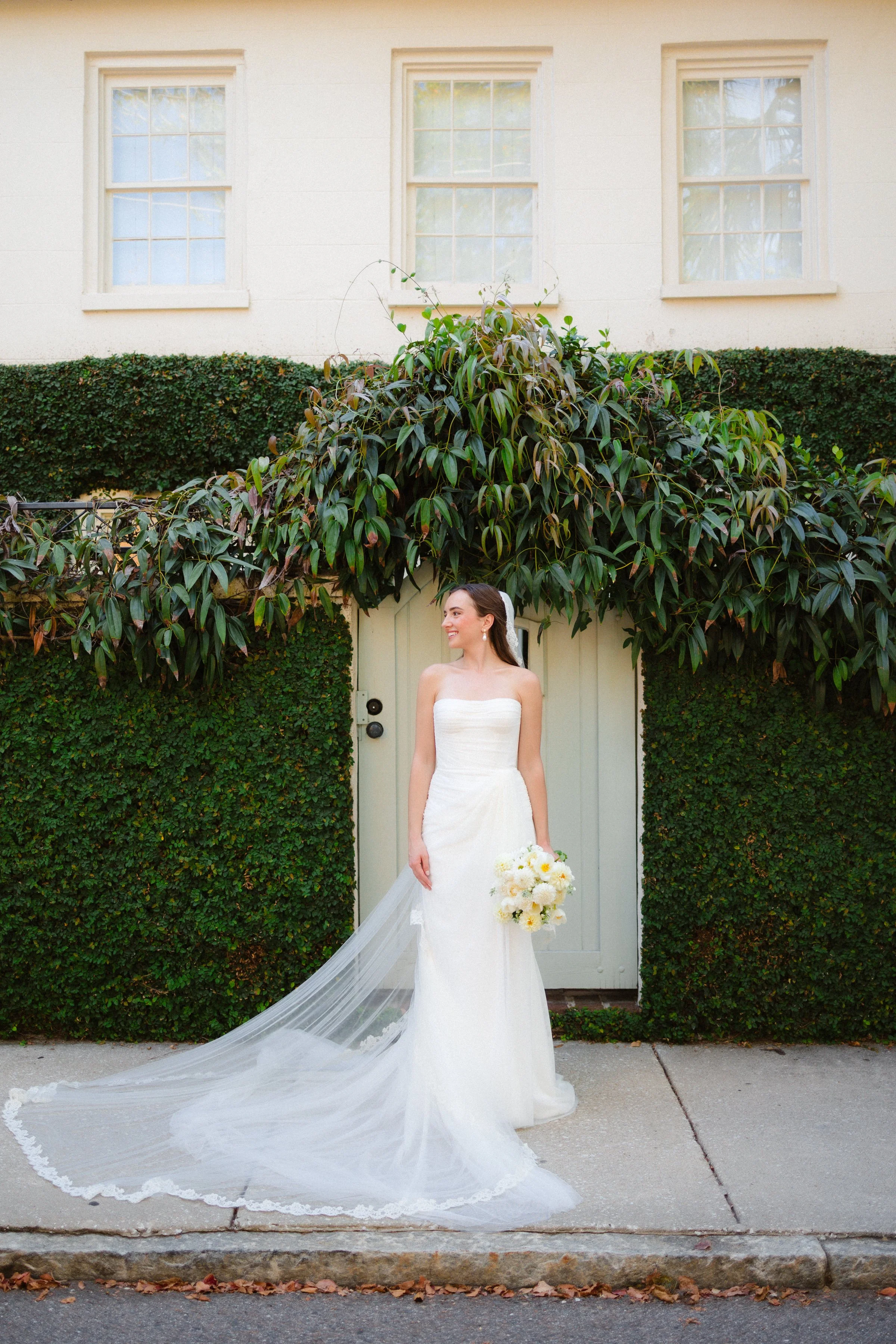 Bride in a white wedding dress holding a bouquet of white flowers standing outside in front of a green hedge and white house with three windows.
