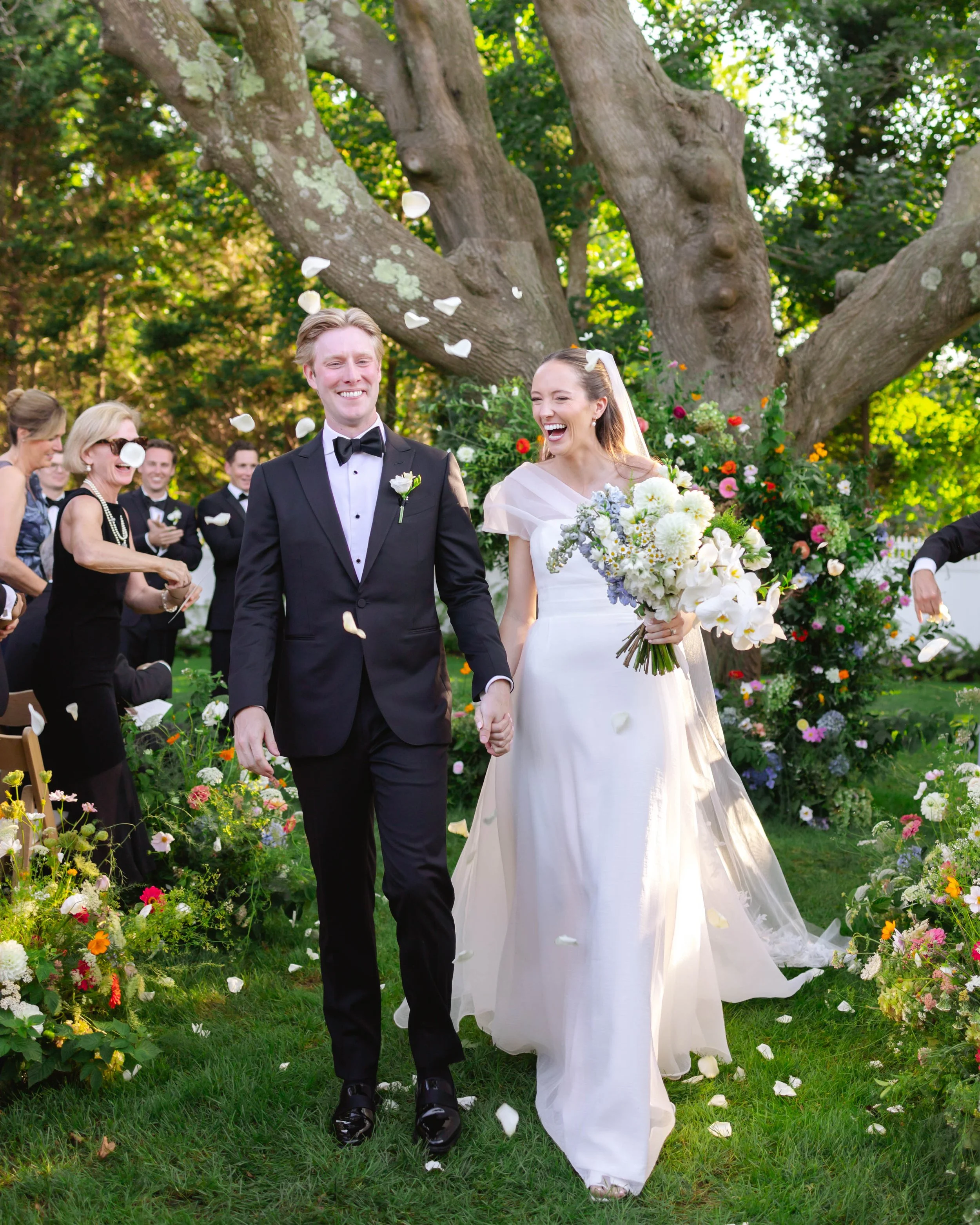 Bride and groom walking hand in hand outdoors under a large tree, smiling, as flower petals are thrown in celebration at their wedding.