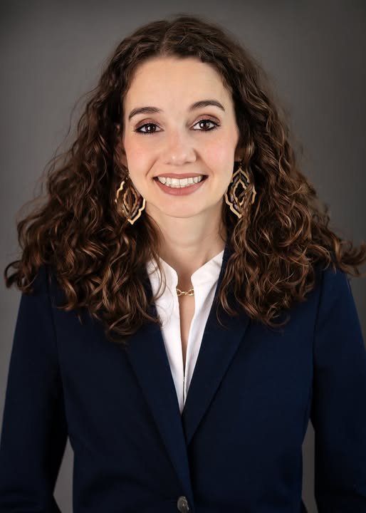 A woman with curly brown hair is smiling, wearing gold earrings, a white shirt, and a navy blazer, against a gray background.