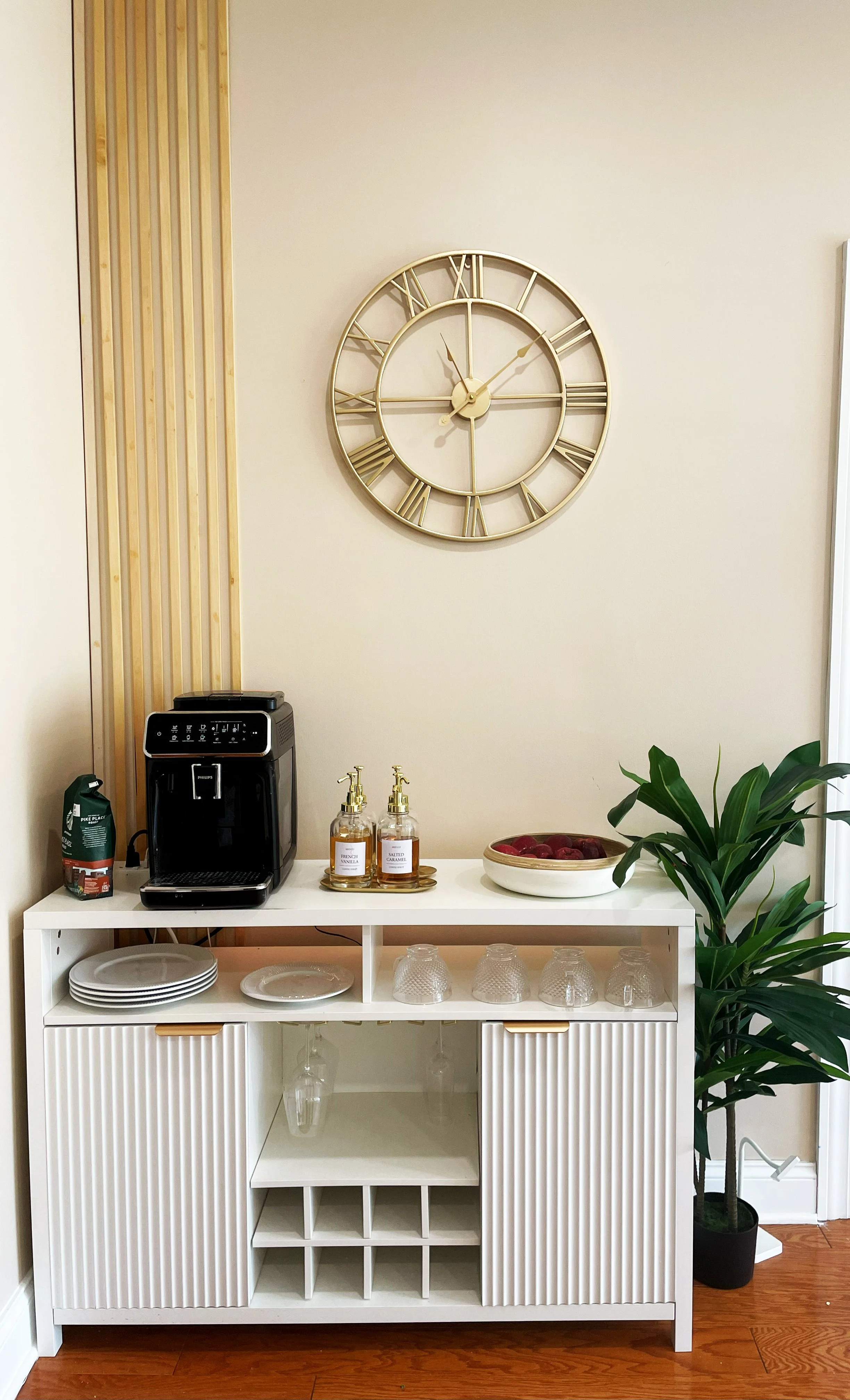 White buffet table holding a coffee maker, soap dispensers, a bowl of red fruit, and glasses. Overhead, a large gold Roman numeral clock on a beige wall, with a wooden slat decor element on the left, and a green potted plant on the right.