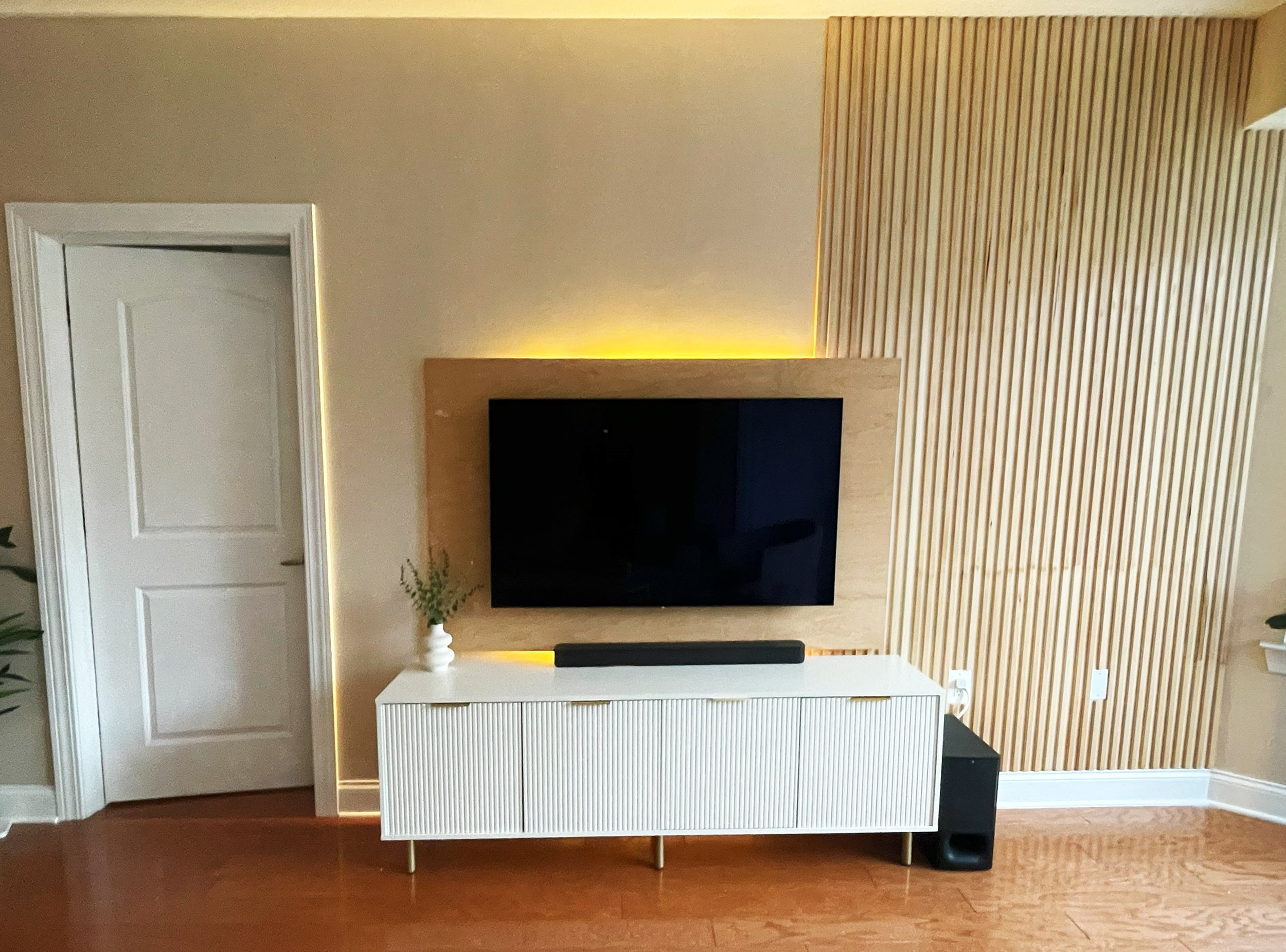 Living room with a wall-mounted flat-screen TV on a wooden panel, white cabinet below with a small plant, and a black soundbar, illuminated with yellow accent lighting.