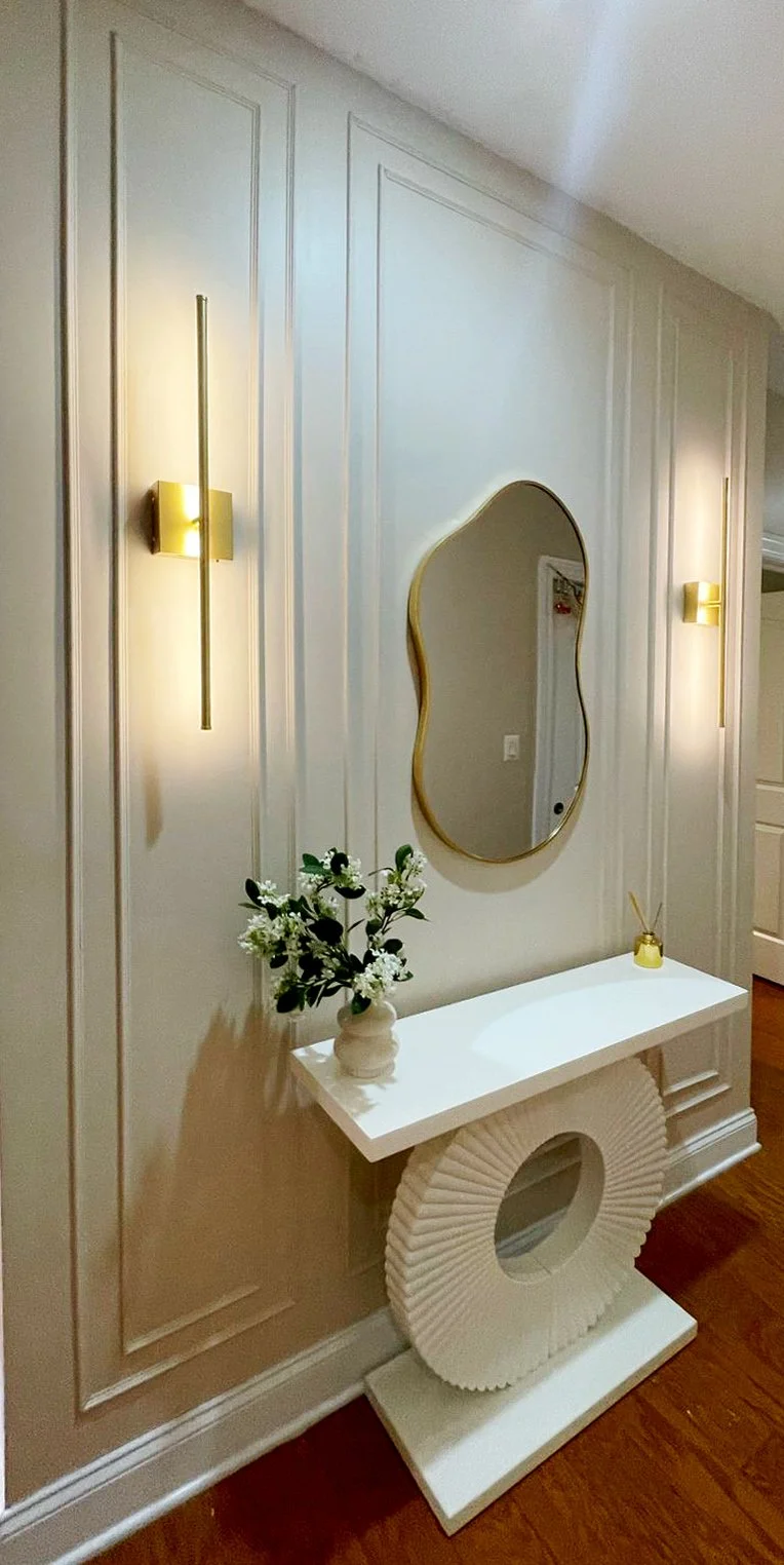 Interior hallway with beige panel walls, two modern wall sconces, a decorative mirror, a white console table with a circular base, a vase with black and white flowers, and a small yellow decorative object.