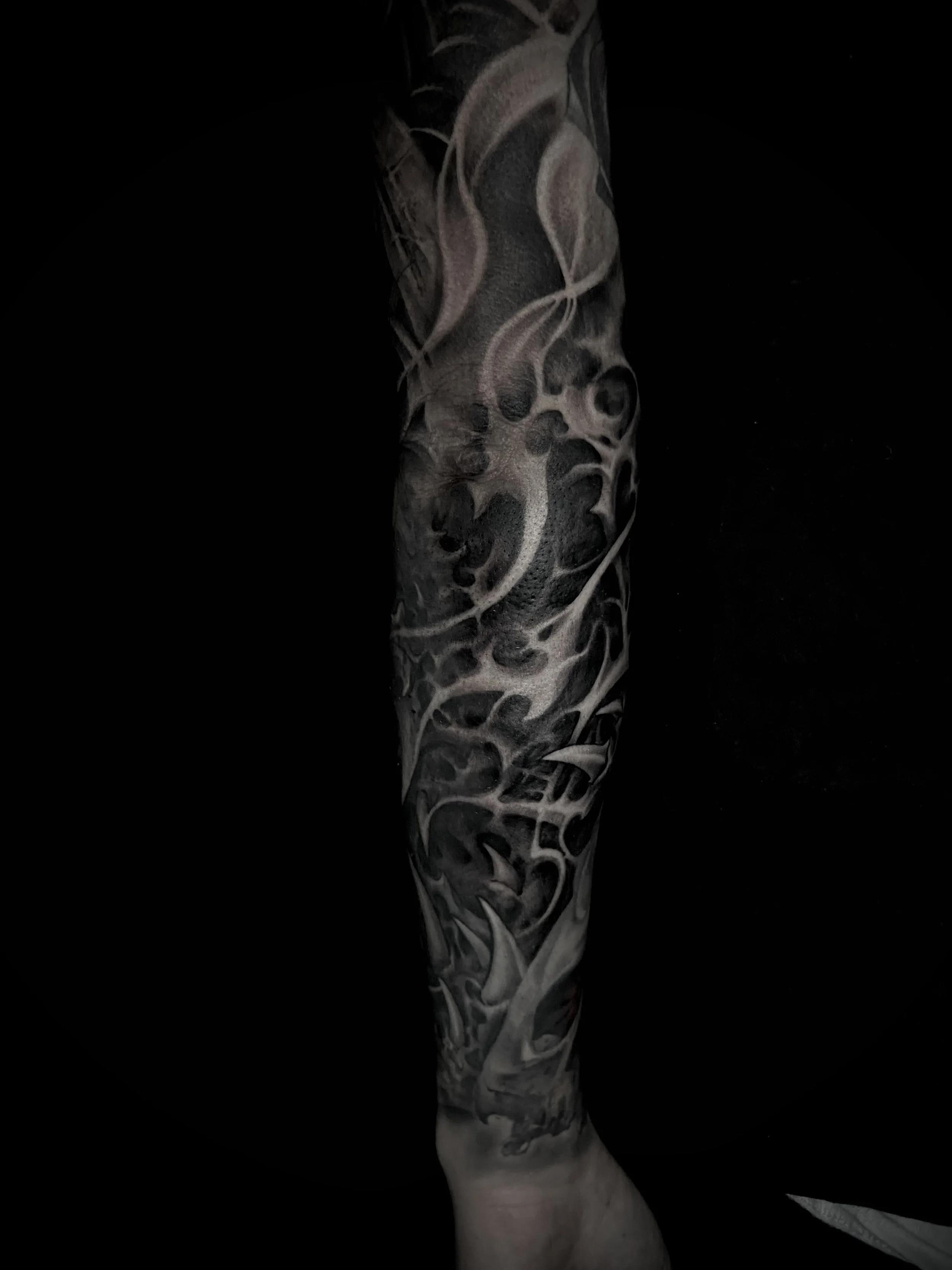 Black and white tattoo sleeve on a person's arm with abstract smoke, skulls, and face elements.
