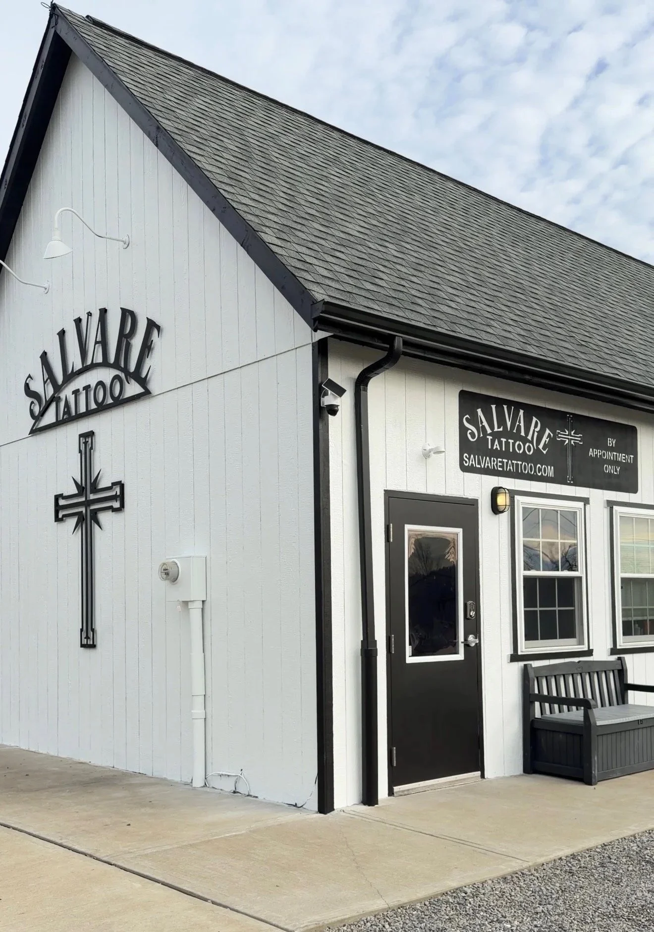 Exterior of a building with signs reading 'Salvare Tattoo' and 'by appointment only'; black door, windows, a bench, and a cross symbol on the wall, under a cloudy sky.