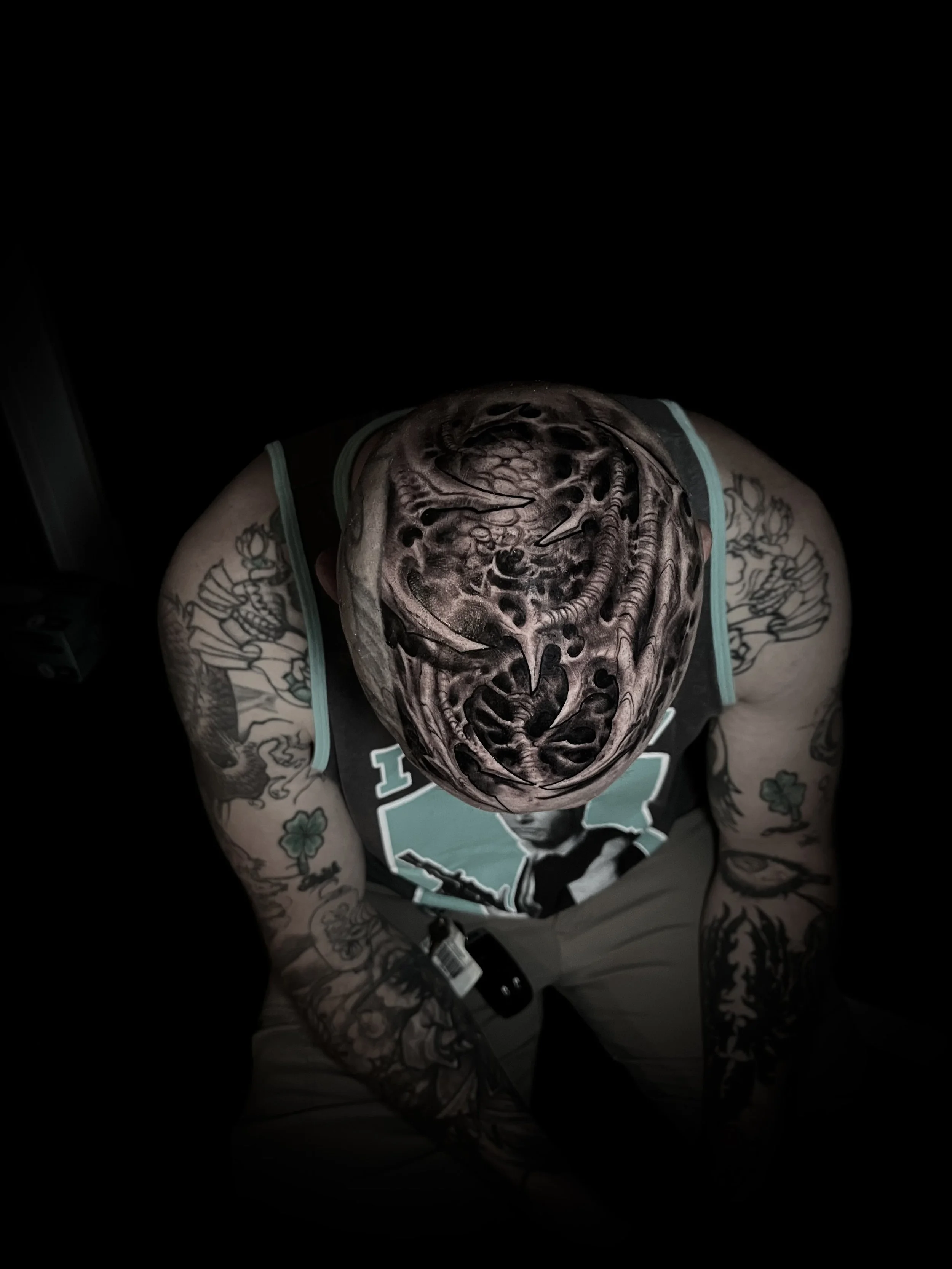 Person with detailed black and grey tattoo sleeves on both arms, wearing a sleeveless shirt, sitting with head bowed down, and wearing a mask or face paint that resembles a creature or monster's face with intricate designs, dark shading, and textures