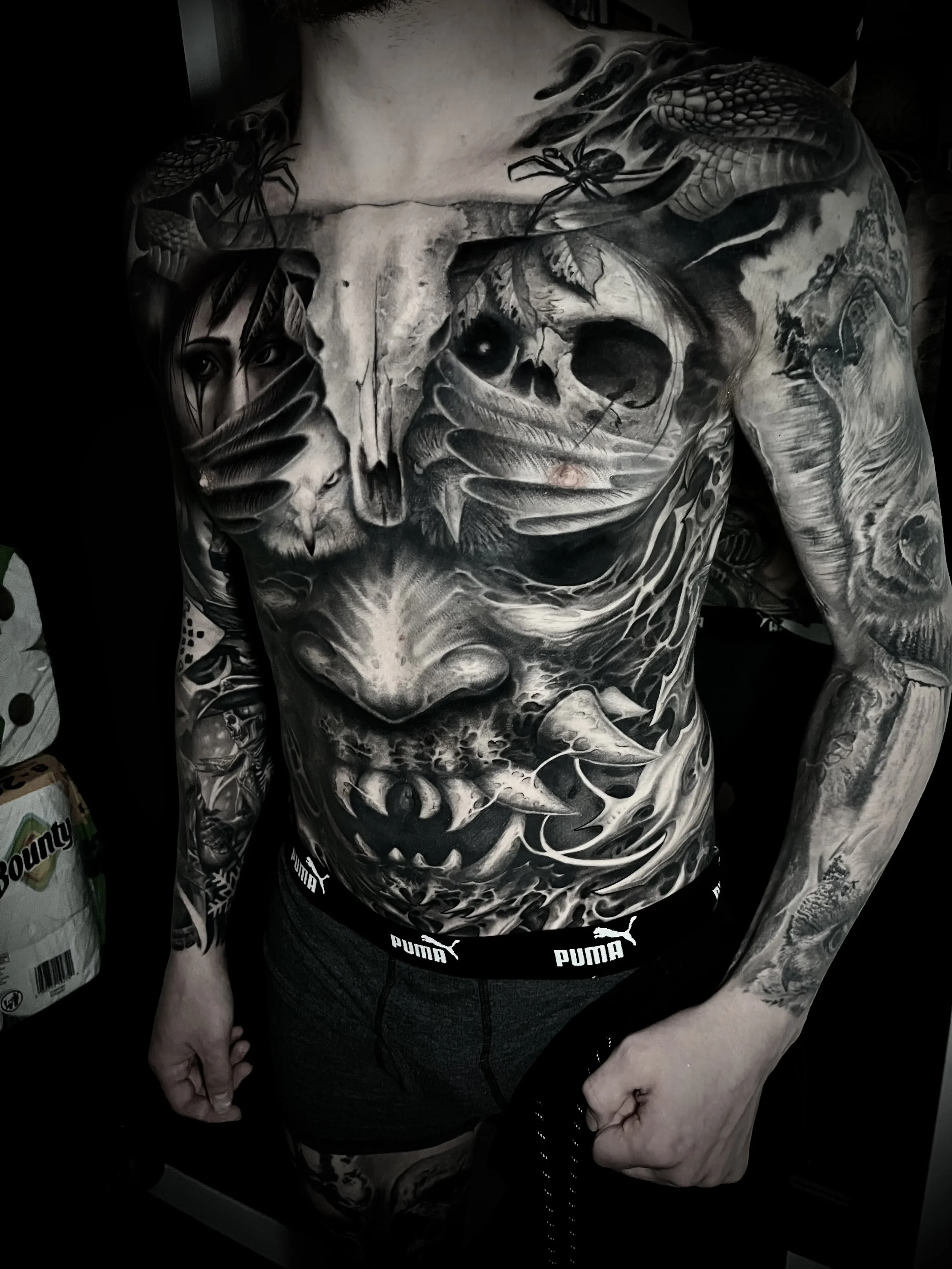 A man with extensive black and gray tattoos covering his chest, arms, and abdomen, featuring skulls, a woman, snakes, and abstract designs.