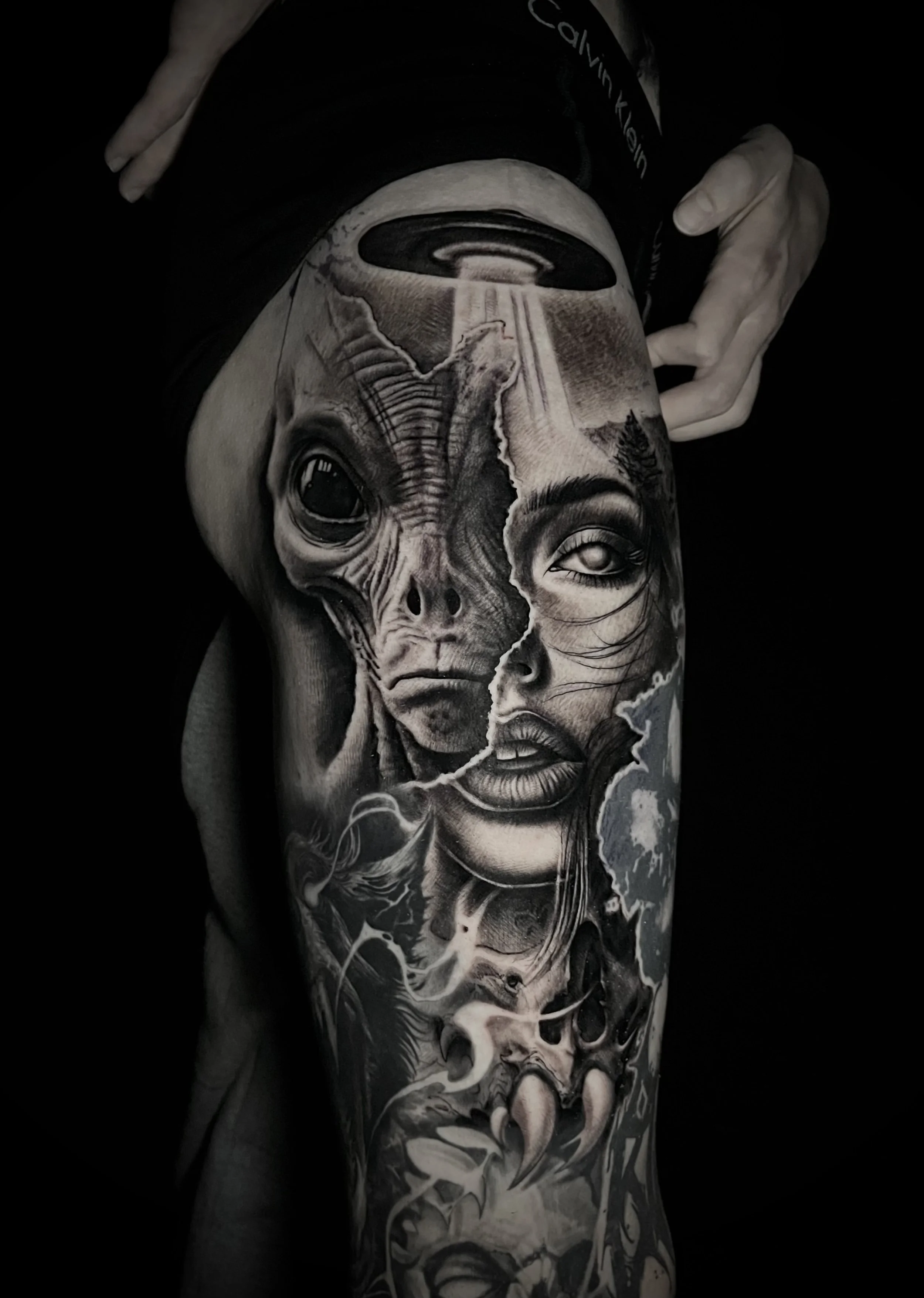 Close-up of a person's arm with a detailed black and gray tattoo depicting an alien and a woman's face, torn and revealing the scene beneath, with an UFO and smoke elements.
