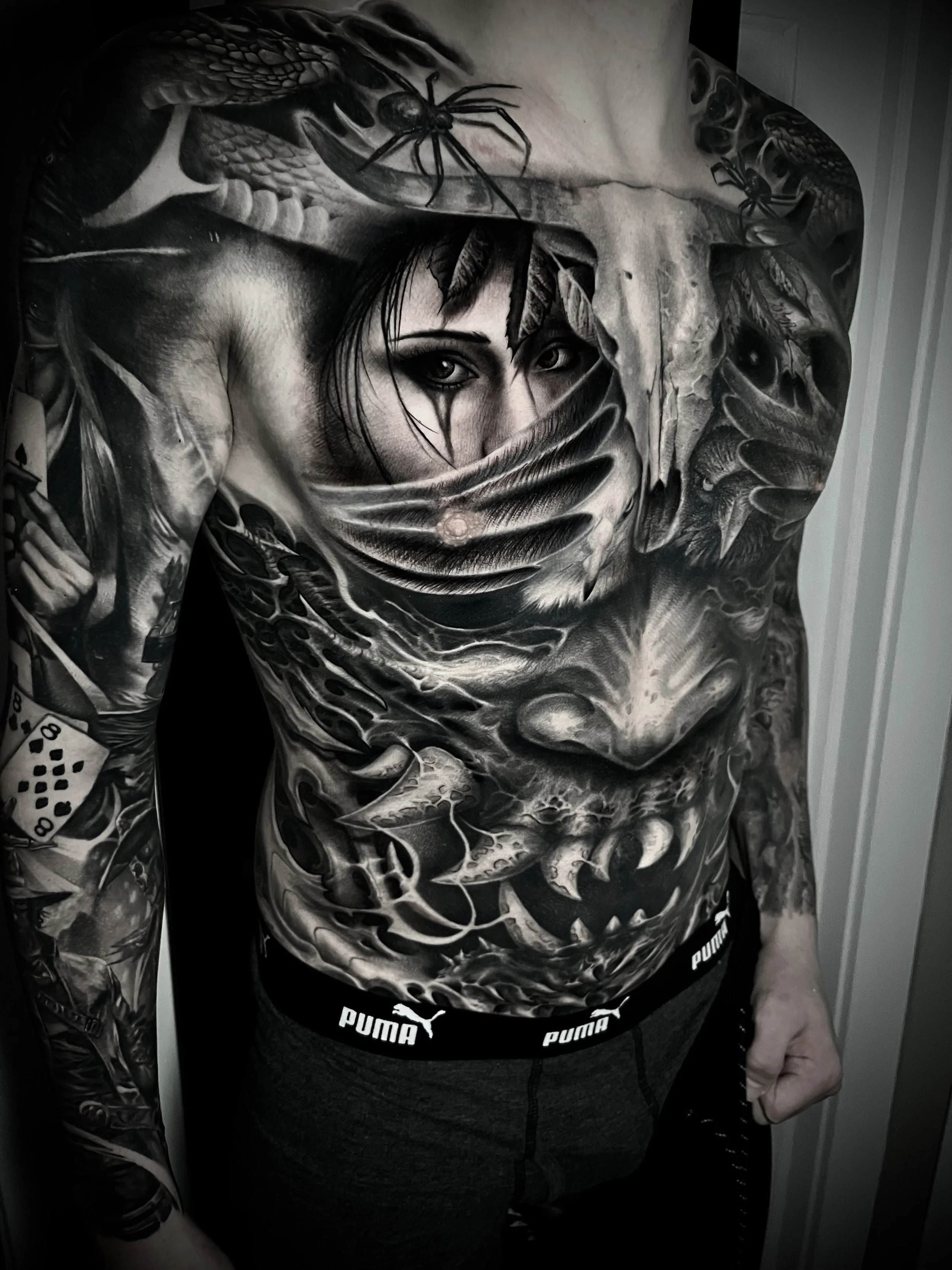 A person's torso and arm covered in detailed black and gray tattoos, including a woman’s face with dark makeup, a snake, and various other intricate designs.