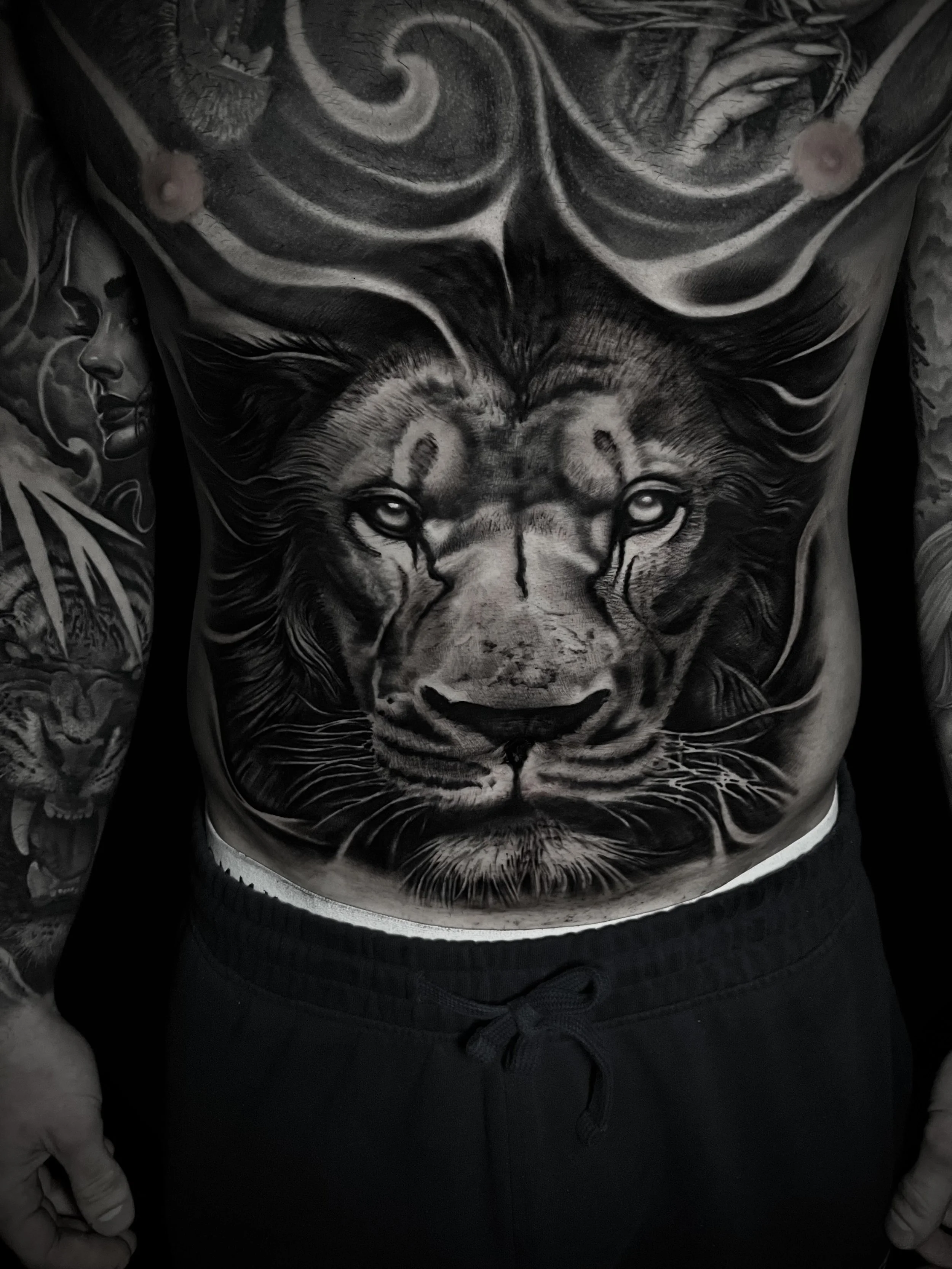A person's chest tattoo featuring a detailed black and gray portrait of a lion with a regal expression, surrounding elements include the face of a woman on the left arm and various artistic designs.