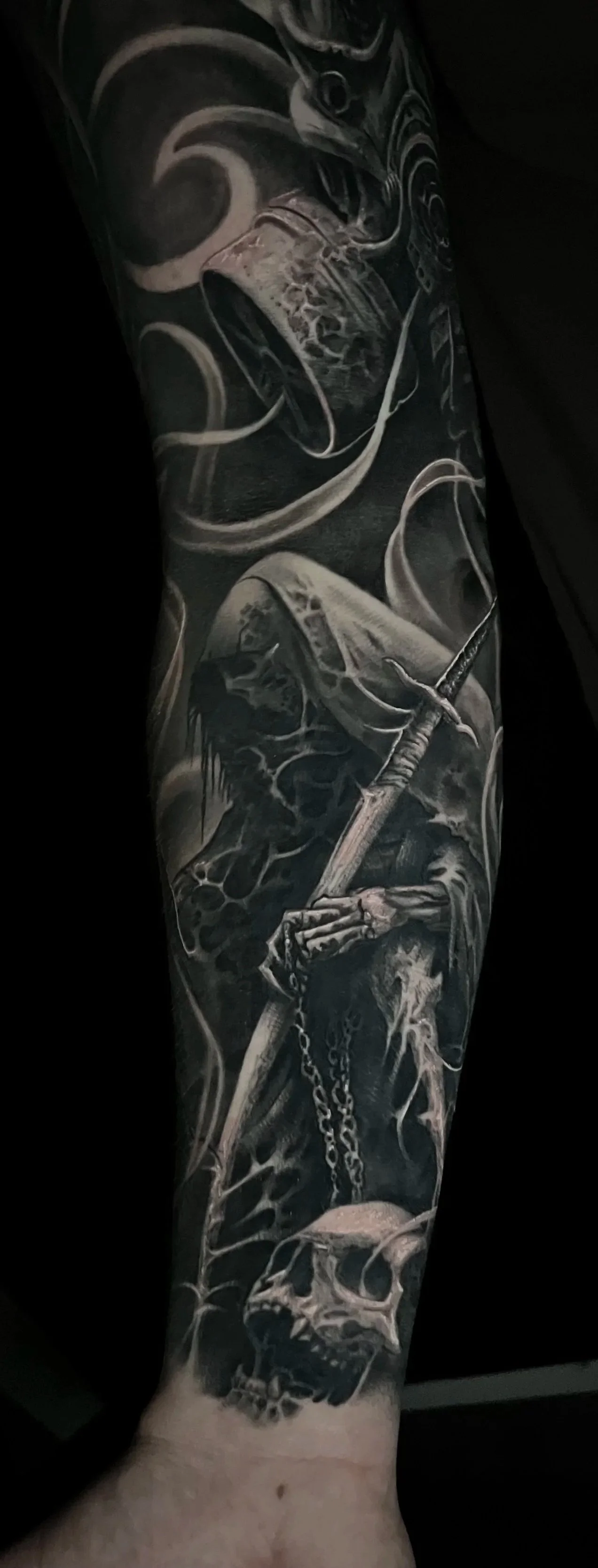 A black and gray sleeve tattoo featuring a skull, a cross with a chain, a hooded figure, and swirling smoke or mist.