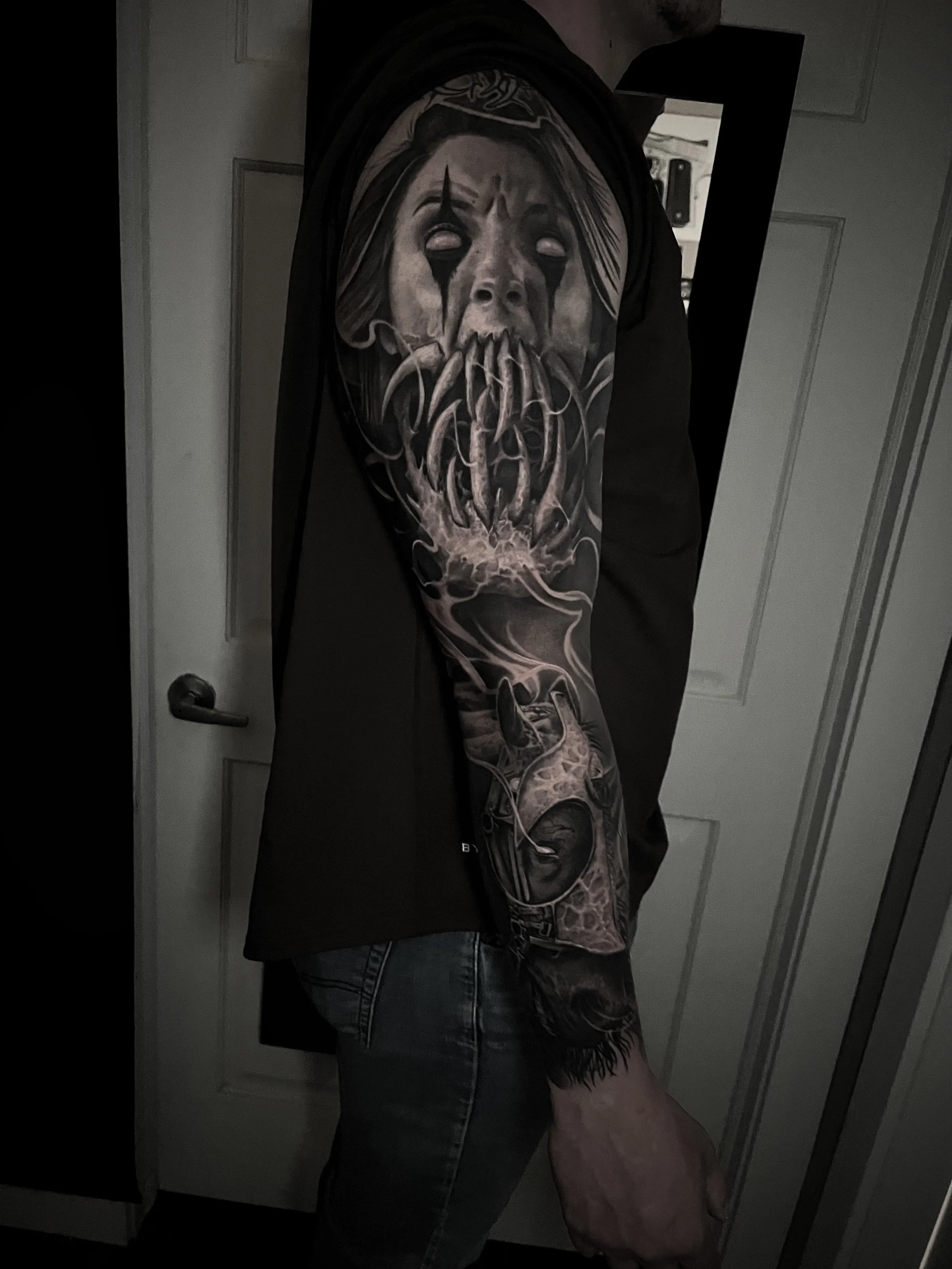Tattooed arm with a detailed black and gray portrait of a woman with tears, tentacles, and smoke elements, wearing a sleeveless shirt and jeans.