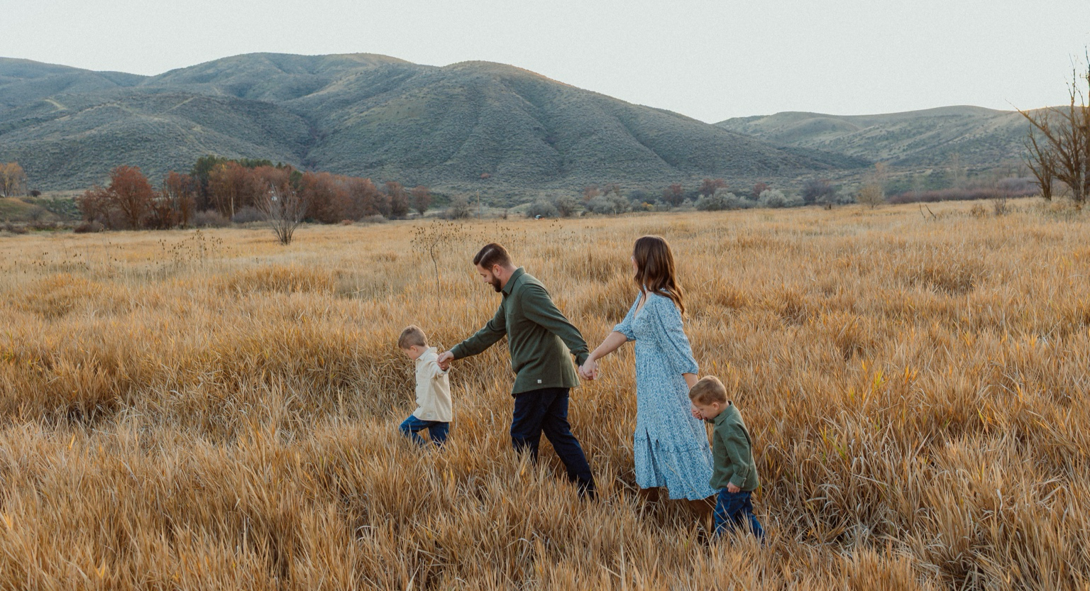 Family of four walking through a field of tall golden grass, holding hands with mountains in the background during autumn.