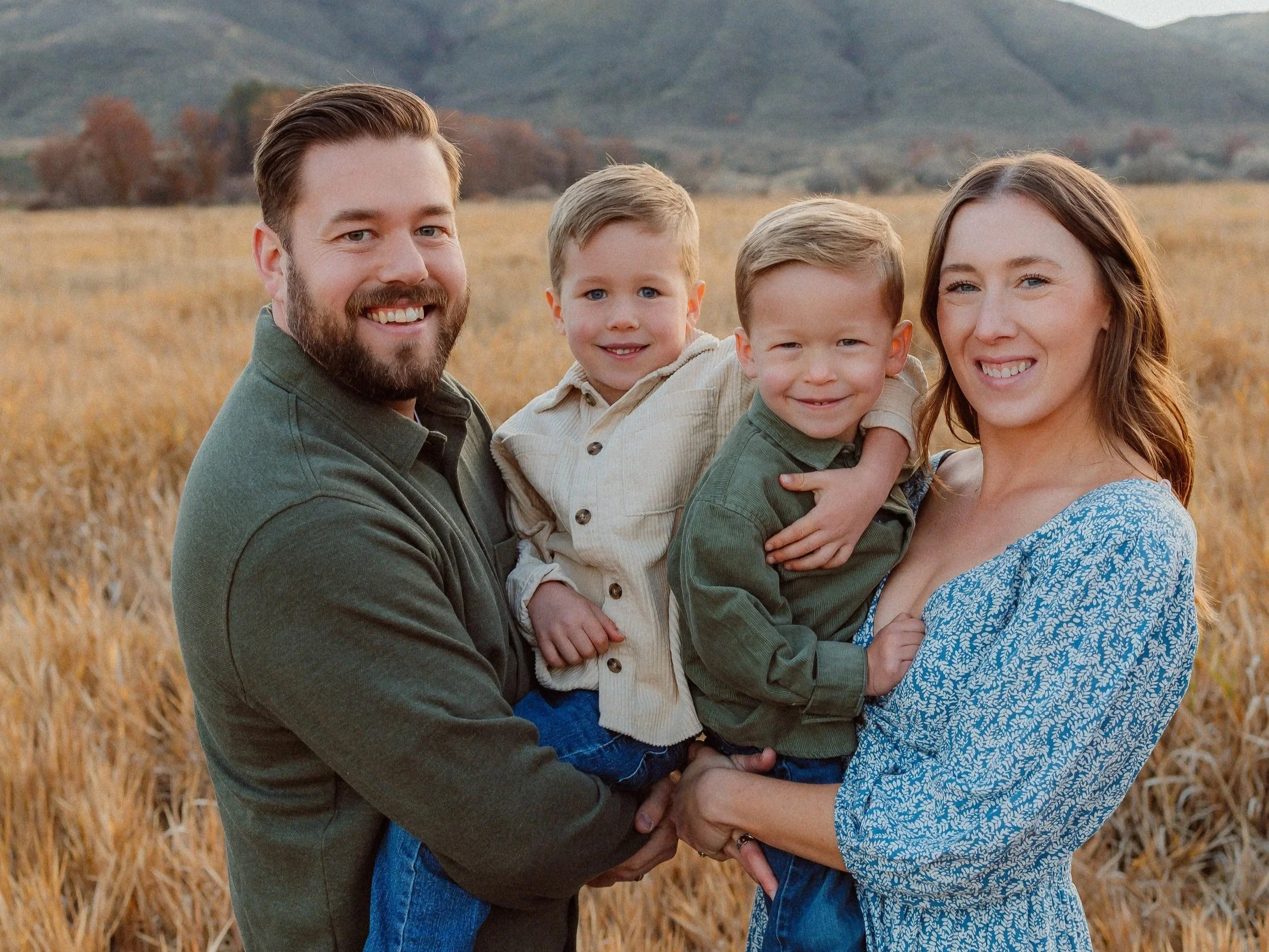 A family of four standing in a field of tall golden grass, with hills and trees in the background, smiling at the camera during sunset.