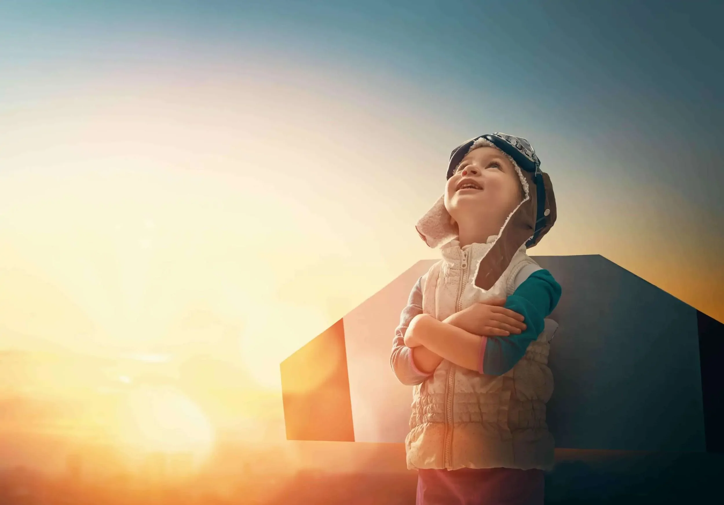 A young child wearing a helmet and a puffy vest, standing outdoors at sunset, looking up with arms crossed, with a large geometric structure in the background.
