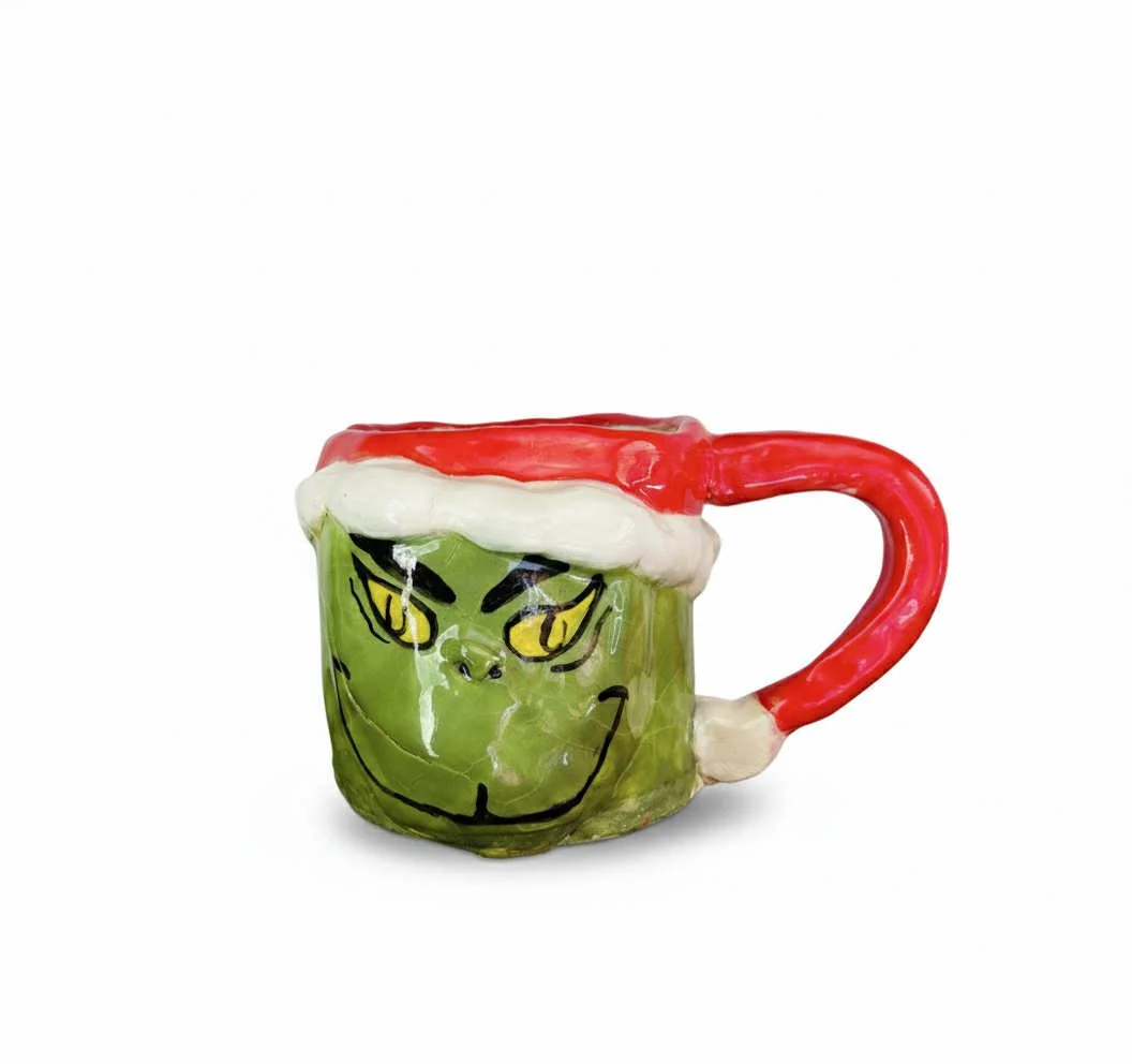 Ceramic mug shaped and painted to resemble the Grinch wearing a Santa hat, with a white band and red top, against a plain white background.
