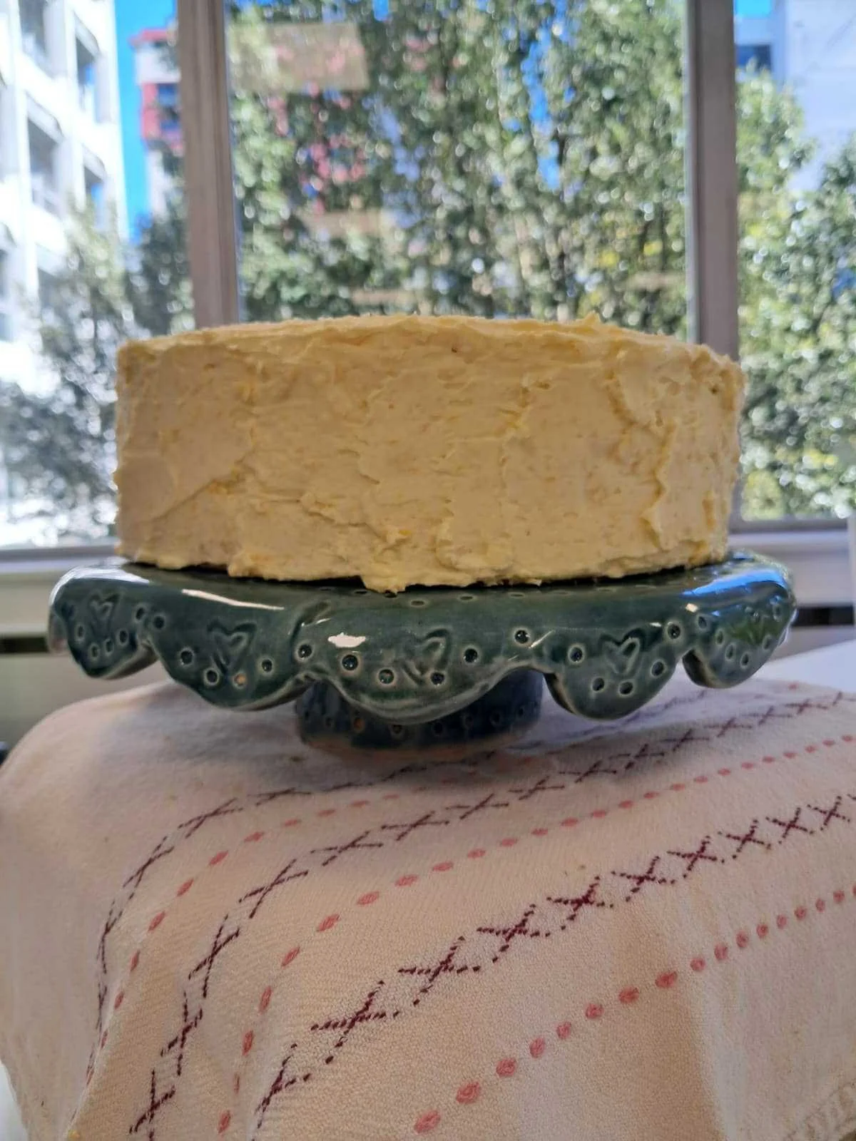 A layered yellow cake on a decorative green ceramic stand, placed on a beige tablecloth with red and black stitched patterns, with a window and trees in the background.