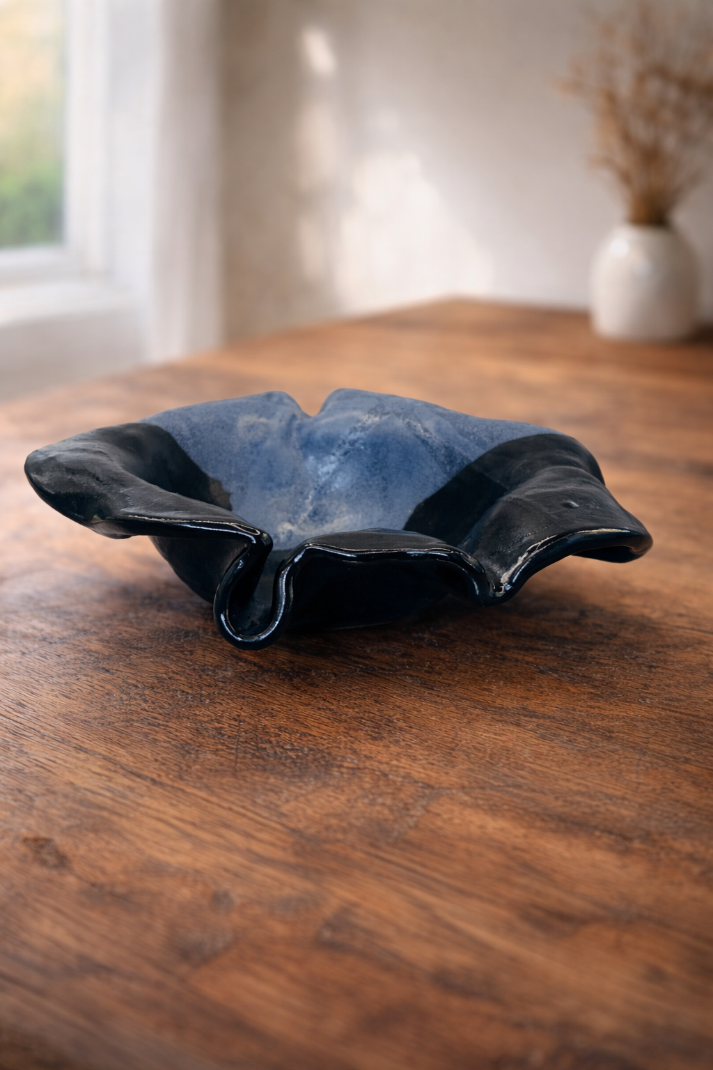 A decorative blue flower ceramic bowl with a wavy, ruffled edge, black on the outside and blue on the inside, placed on a wooden table in a well-lit room.