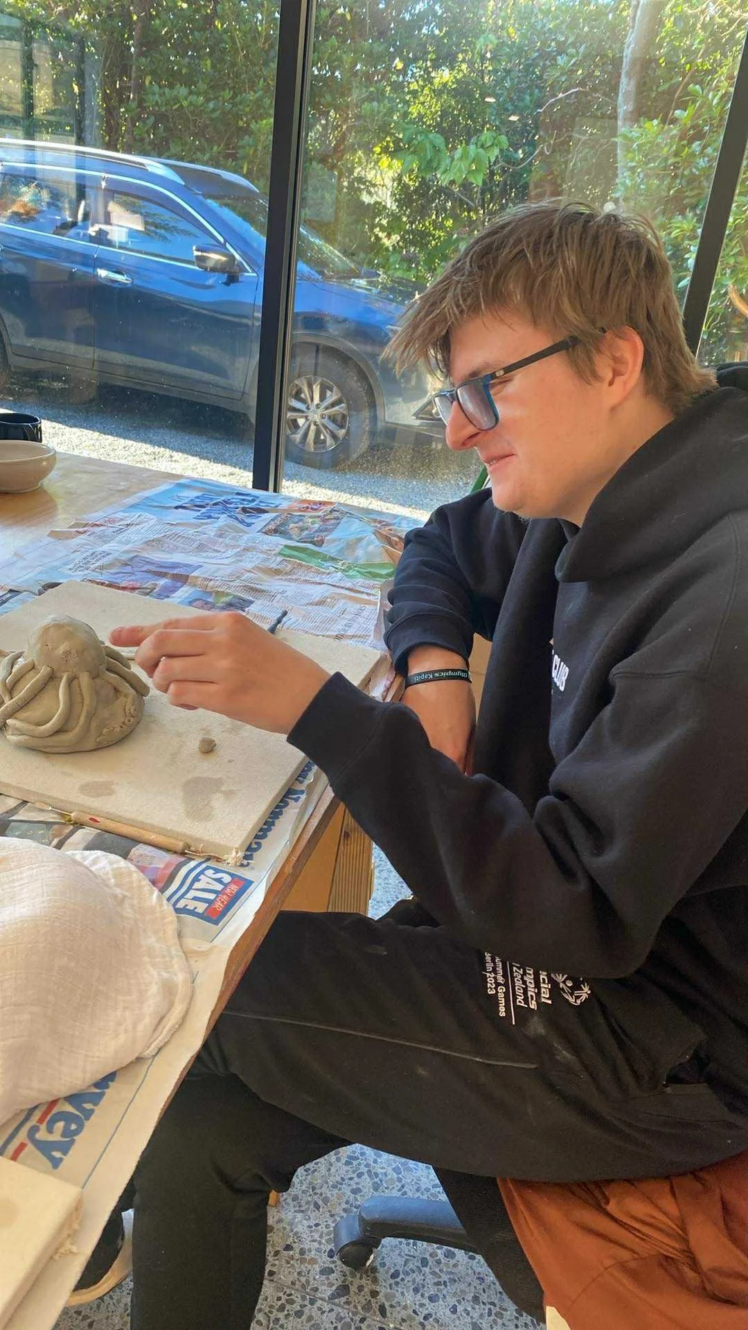 Louis with glasses and a black hoodie sitting at a table, engaged in sculpting clay. He is working on a clay octopus. The table is covered with newspaper and paper towels, with a large window behind him showing a parked blue car and green foliage.