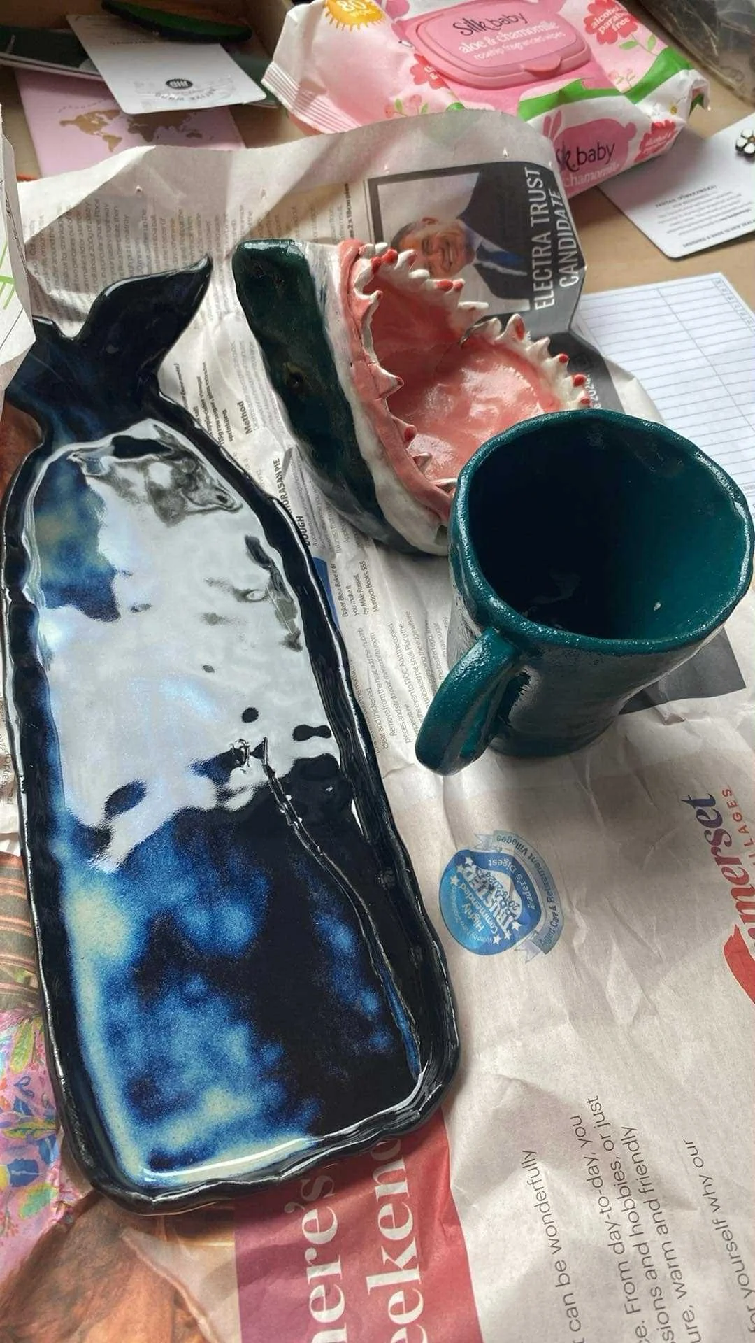 Decorative ceramic items including a shark-shaped tray, a large tooth-shaped dish, and a mug, placed on newspaper with a pack of baby wipes and papers nearby.