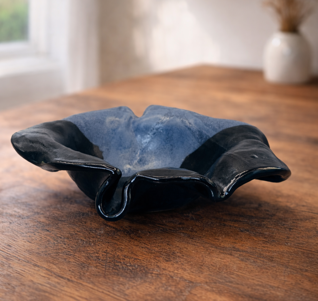 A decorative blue flower ceramic bowl with a wavy, ruffled edge, black on the outside and blue on the inside, placed on a wooden table in a well-lit room.