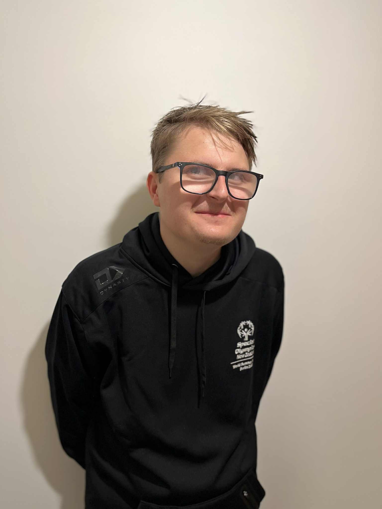 Louis with glasses and short, light brown hair, wearing a black hoodie with a Special Olympics logo on the chest, standing against a plain white wall.