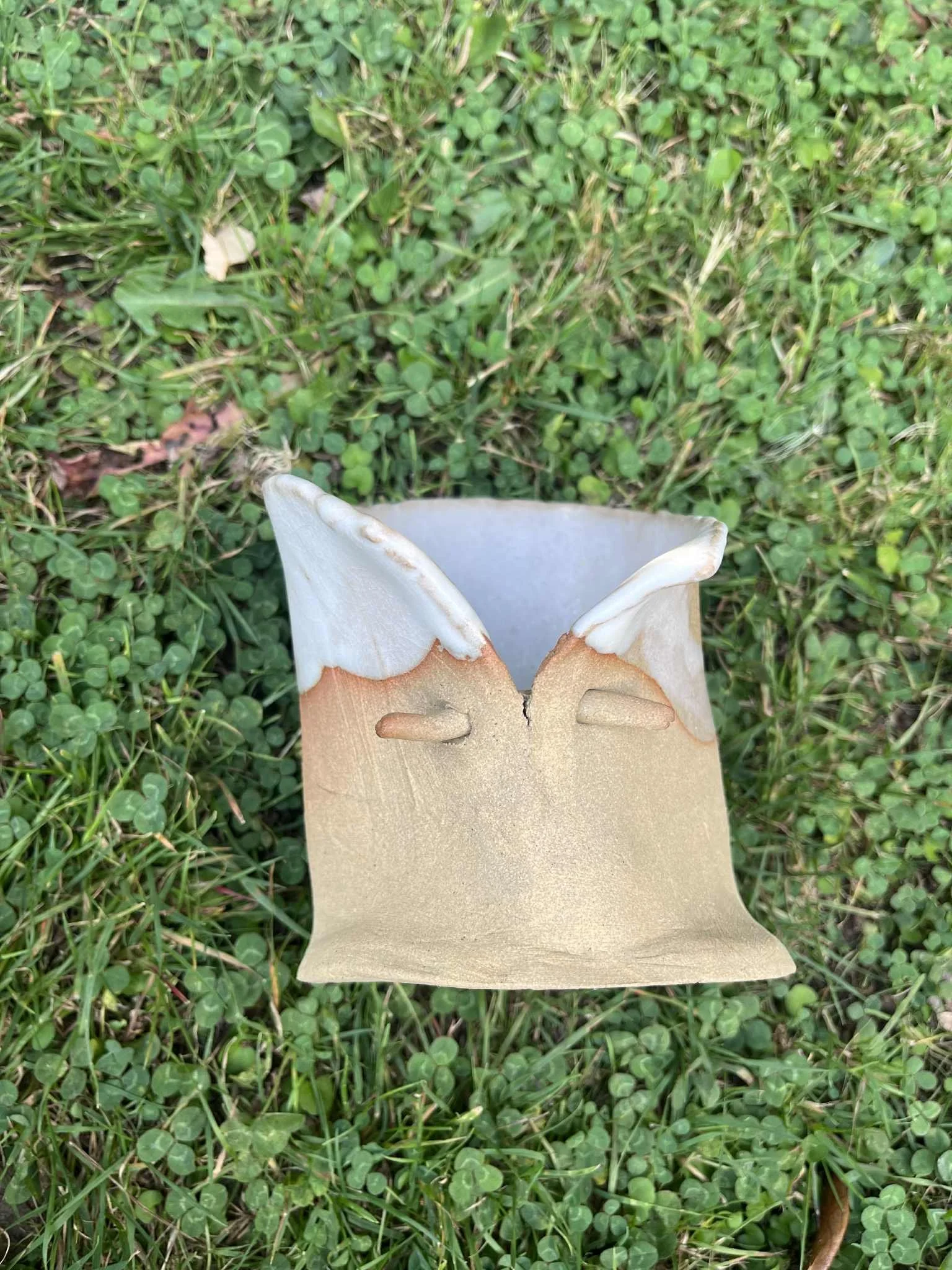 A ceramic mug lying on grass with  a butterfly shaped opening at the top.