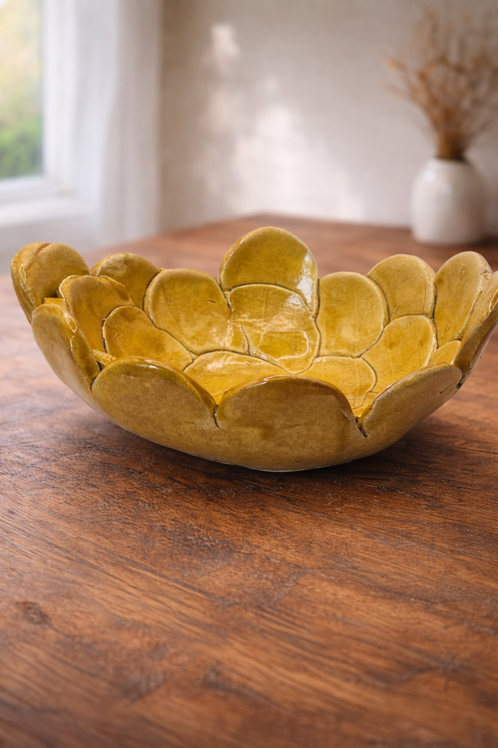A decorative yellow ceramic bowl with a flower petal shape, glazed in yellow, placed on a wooden table with a blurred indoor background.
