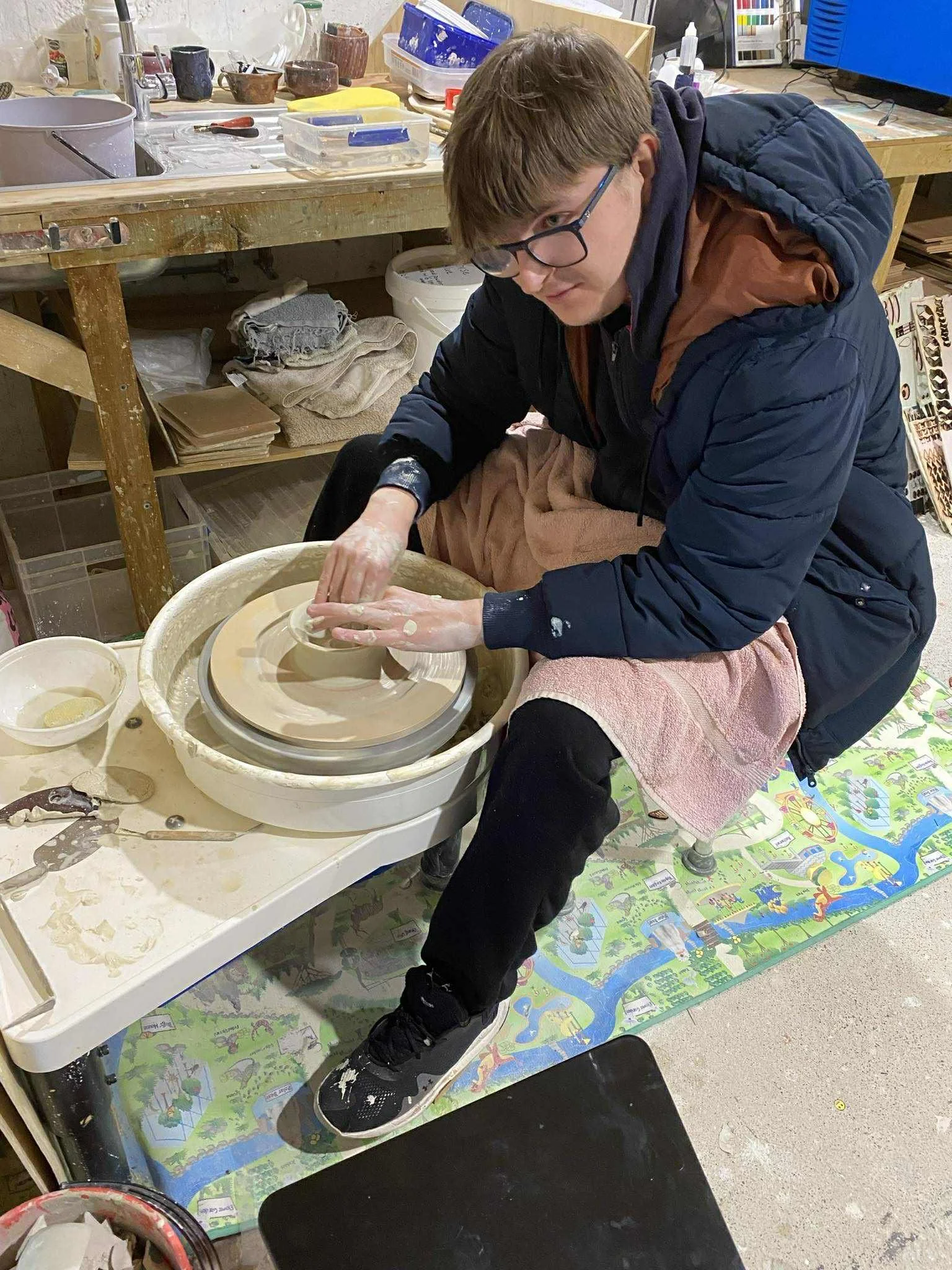 Louis with glasses and brown hair working on a pottery wheel, shaping clay with his hands in a cluttered workshop.