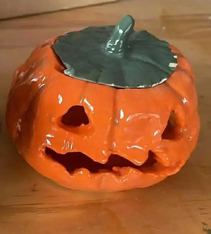 A ceramic pumpkin with a carved face and a green stem on top, sitting on a wooden surface.