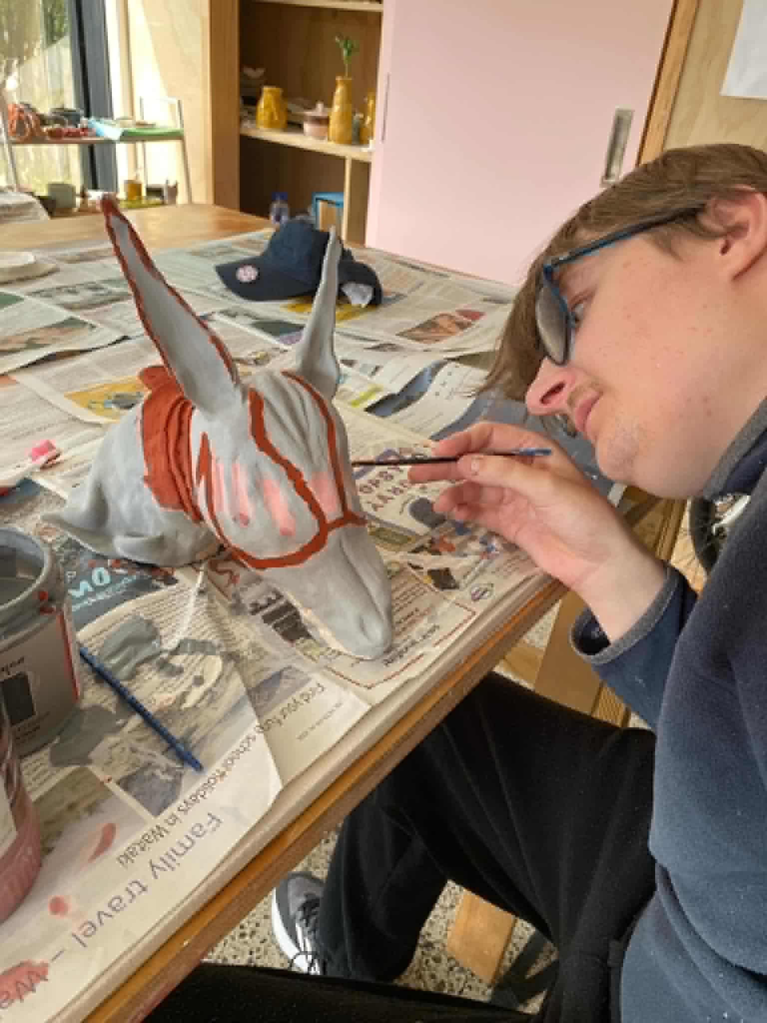 Louis with glasses painting a ceramic figure with pointy ears and with orange details at a cluttered table covered with newspapers in a room with shelves and a window.