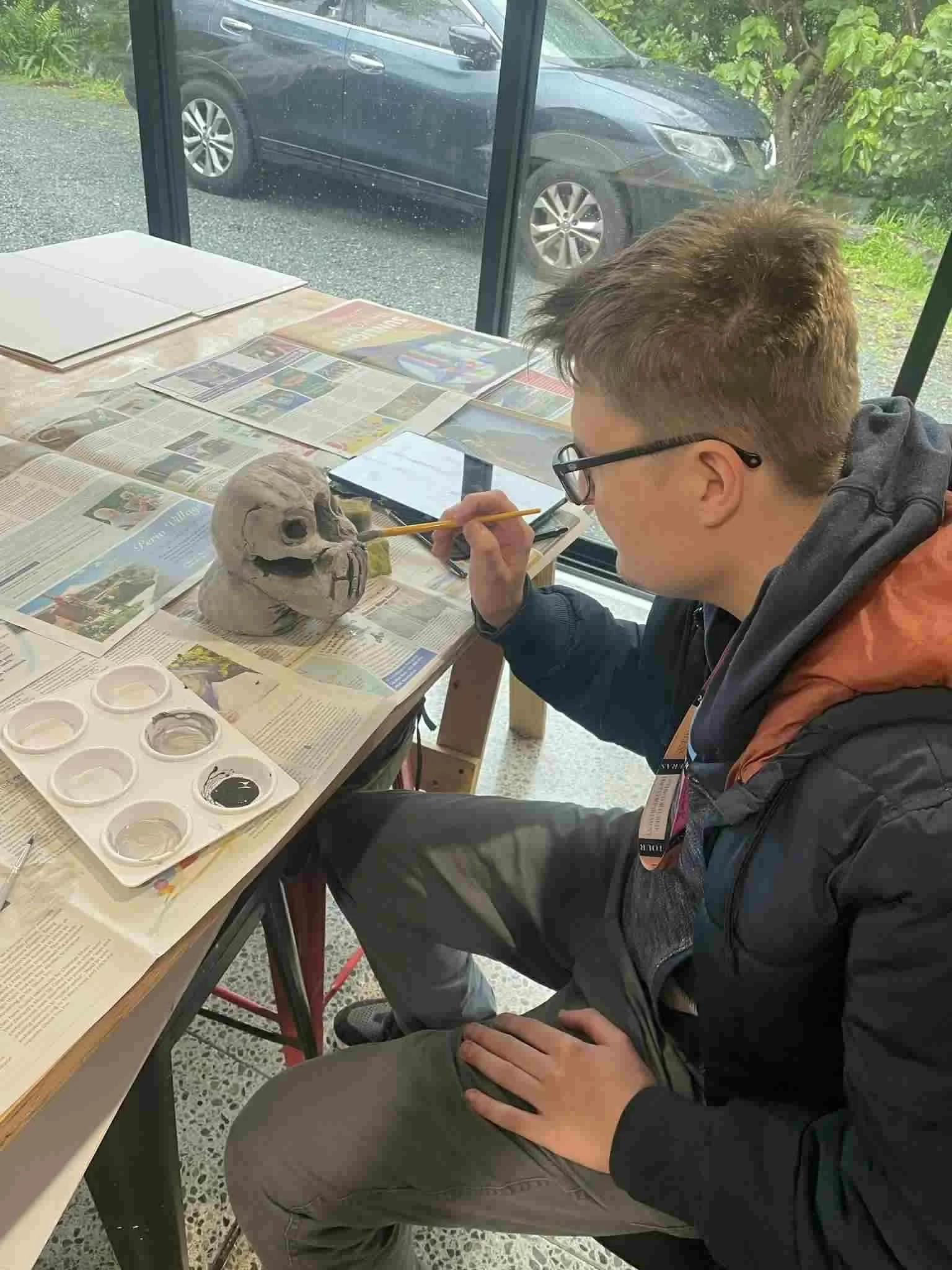 Louis wearing glasses painting a Dr Who skull sculpture at a table covered with newspapers and paint palettes, with a car parked outside visible through a large window.