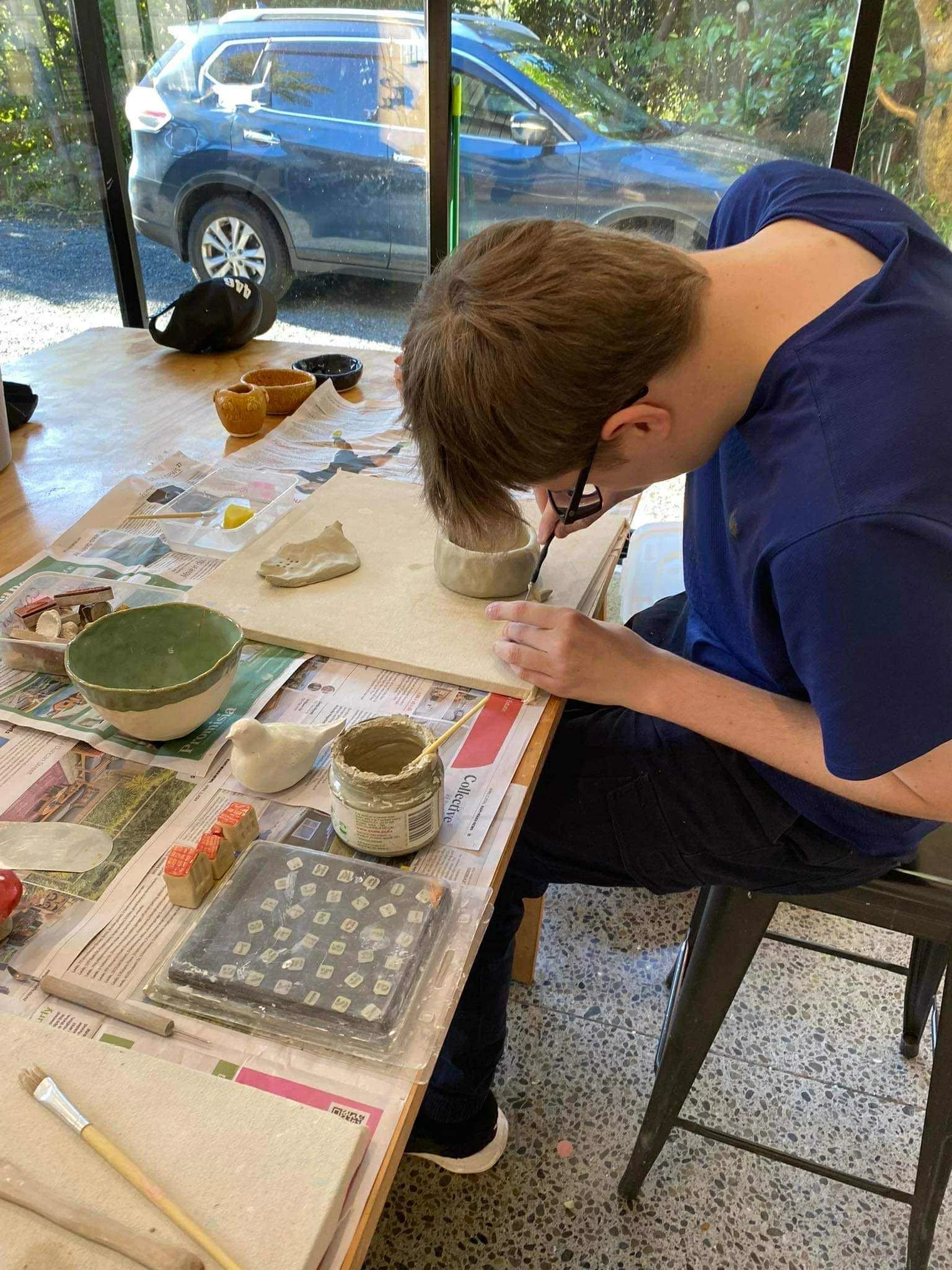 Louis working on a pottery project at a table with various clay and ceramic tools, surrounded by newspapers, with a window showing a parked car outside.
